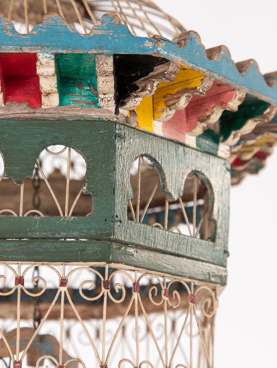 An ornate vintage polychrome wire birdcage featuring a yellow door and two towers. Wooden molding painted in brilliant colors surrounds the crest of the lower level and the taller tower. Each tower features one swing. The wire is shaped to mimic a