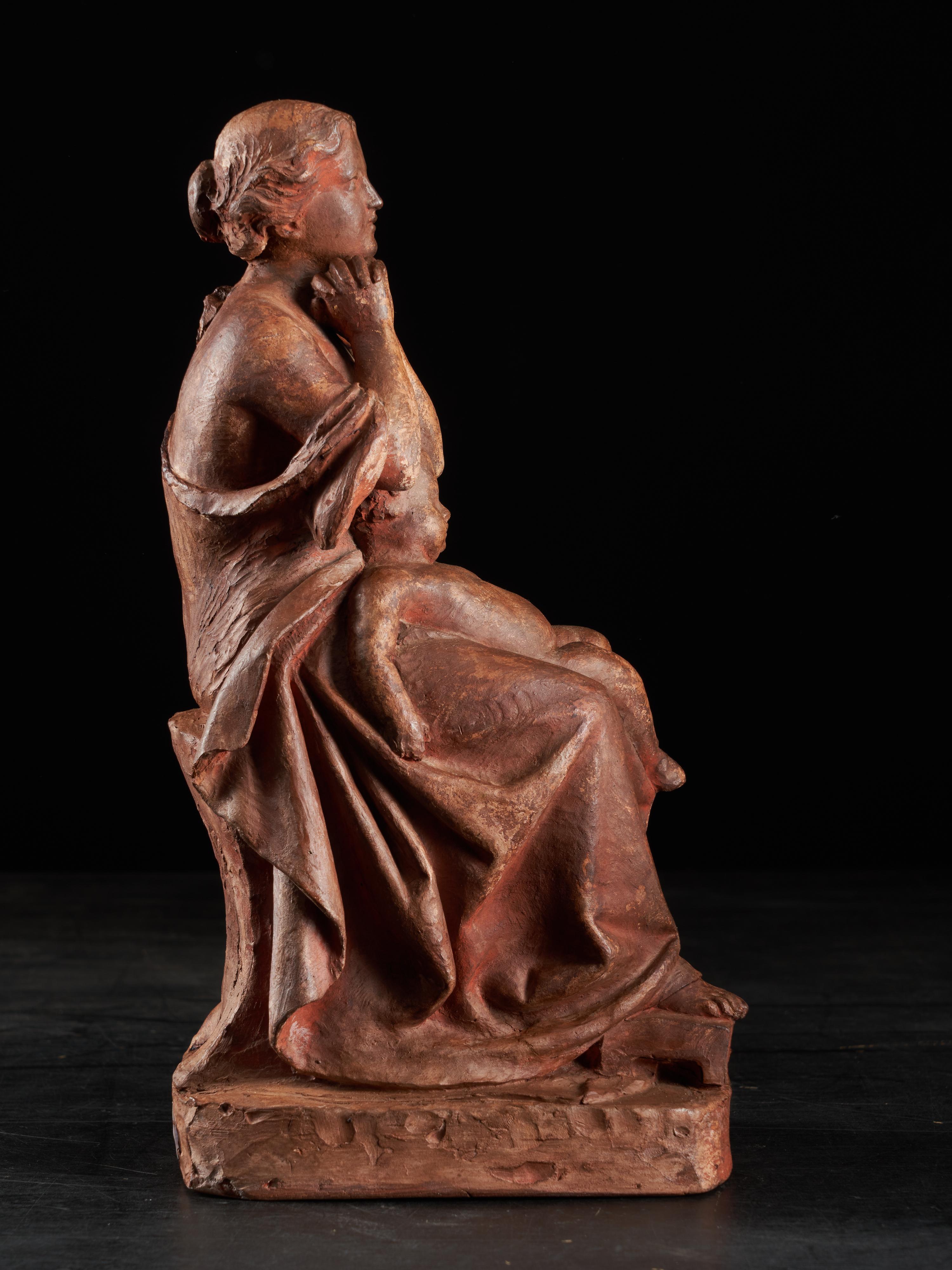 A lovely small terracotta statue of a young woman and a child. The woman is seated on a stool and has her hands clasped together. The young boy is sleeping on her lap with his limbs spread wide open. The sculpture shows the pleads and hair creases