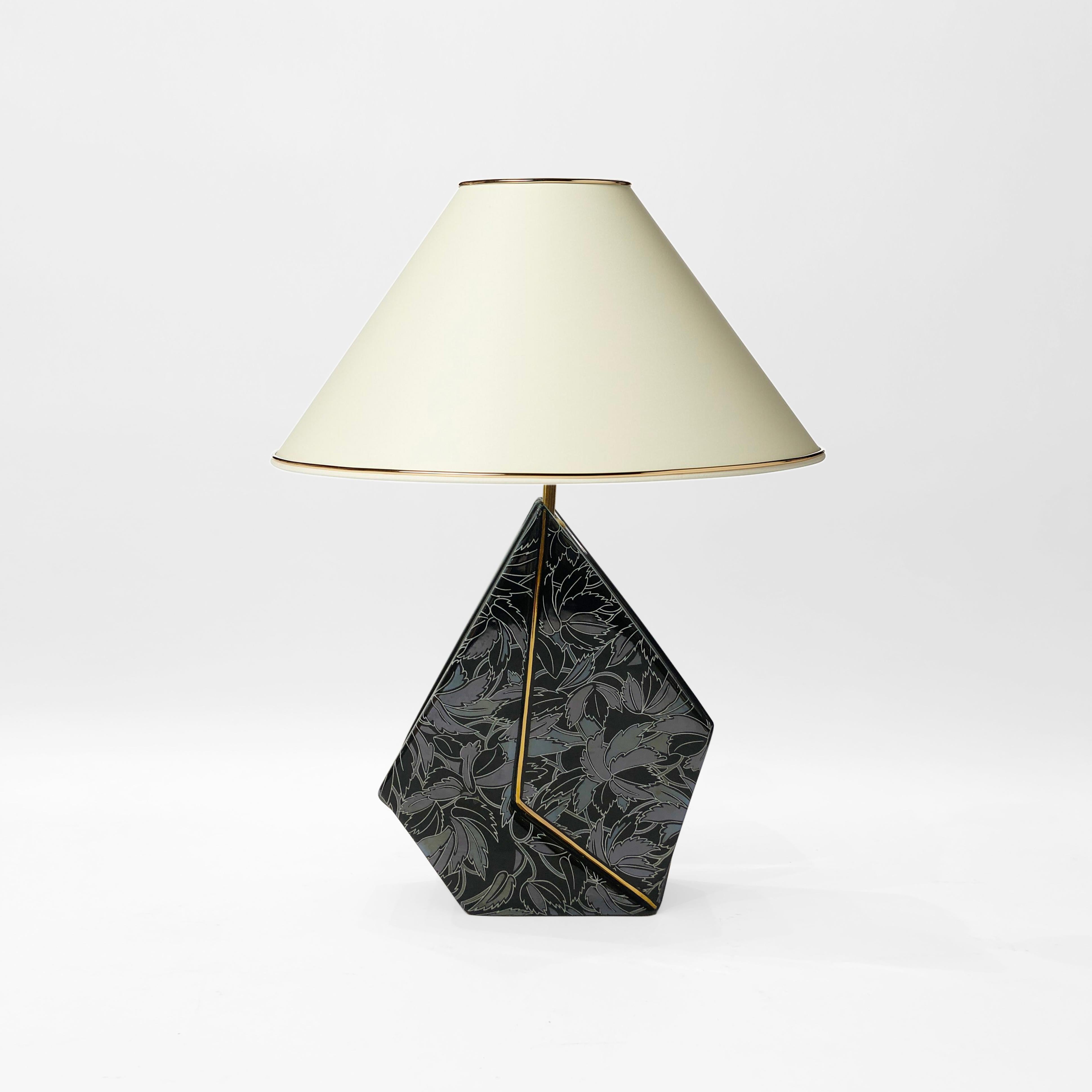 A geometric table lamp, made of glazed ceramic, and finished with a leaf motif. An irregular pentagon, this piece features a jutting brass strip running through the middle to the bulb fitting, creating a distinct left and right to the base. Each