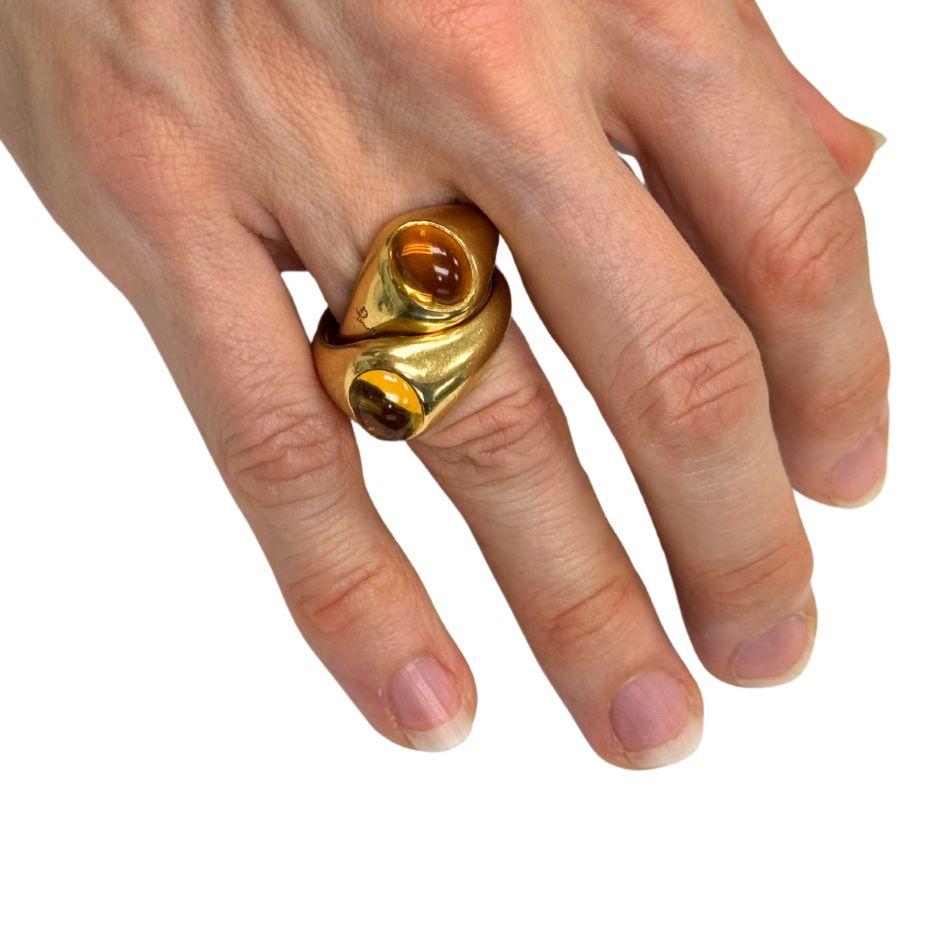 This Pomellato ring features a unique design composed of two interwoven 18k yellow gold bands, each set with a cabochon-cut citrine gemstone. Circa 1990s, the ring showcases the brand's signature blend of sculptural form and rich color. When worn