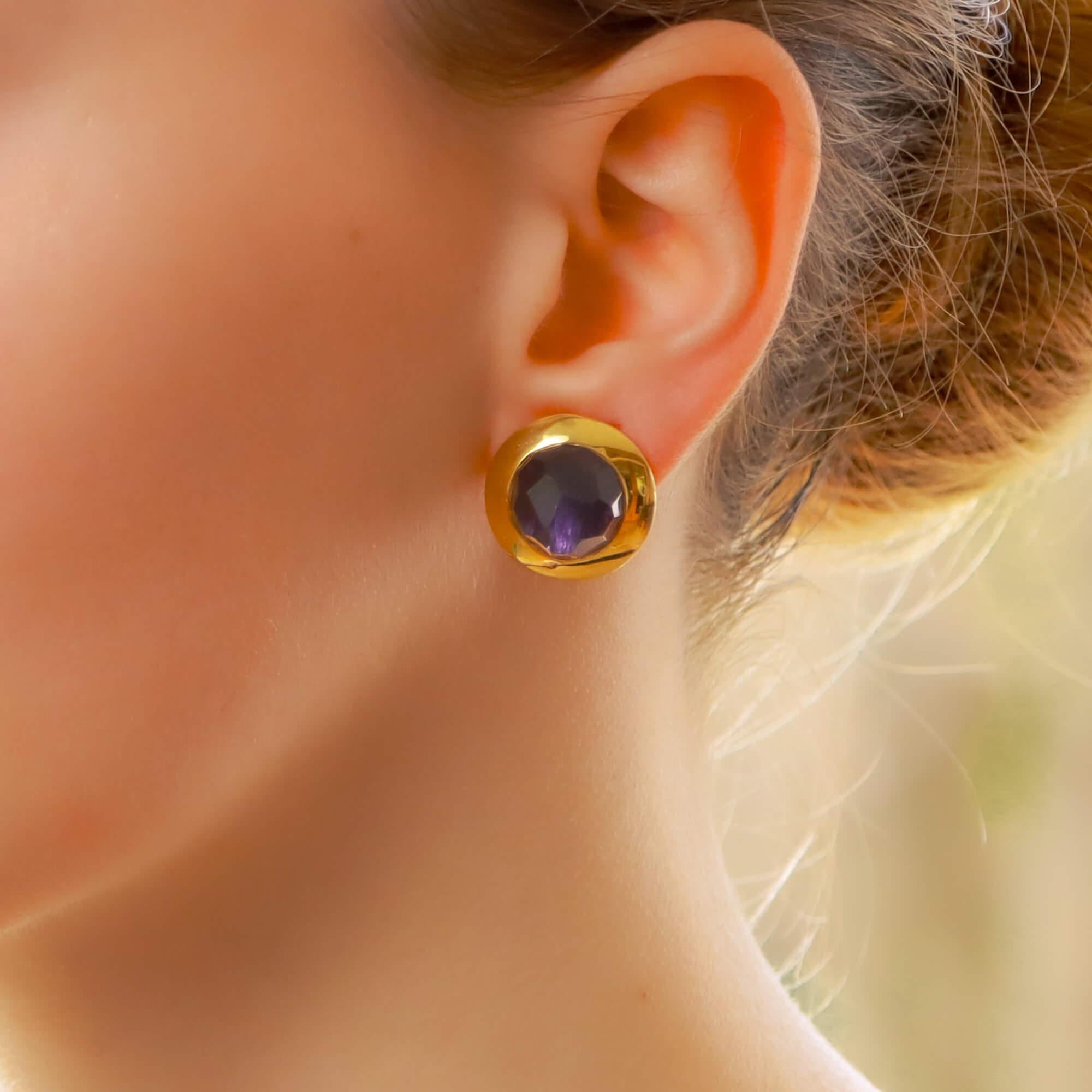 A stunning pair of signed Pomellato purple iolite clip earrings set in 18ct yellow gold. Each earring depicts a shield and features a circular faceted dome iolite of a purple colour. 

This iolite is rubover-set into an open-back polished yellow
