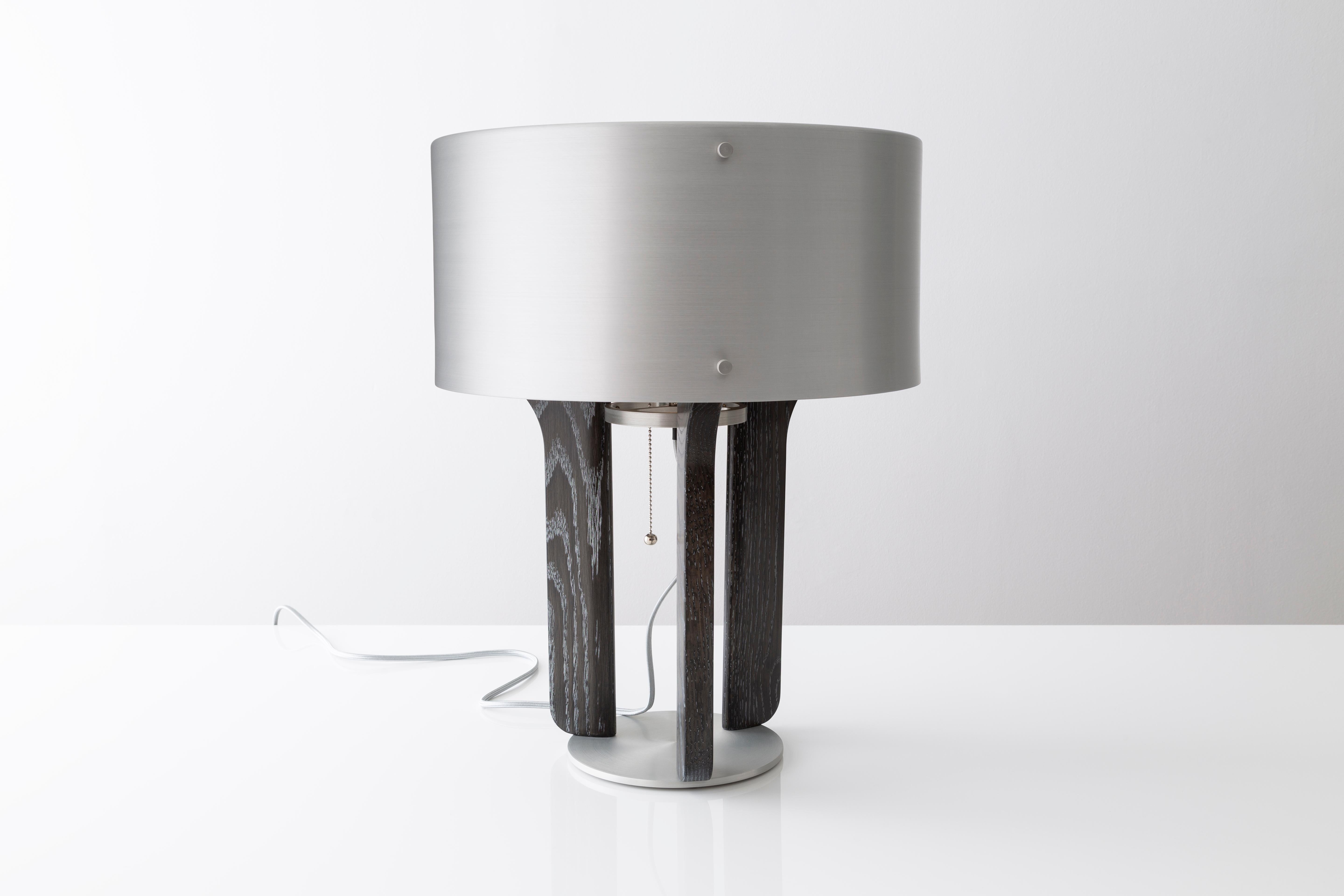 The Pommer table lamp features a satin anodized drum shade with a trumpet like interior. Warm white light radiates light from its core. LED bulbs are supplied and incandescent can be used as well. The legs of the lamp are carved from solid wood and