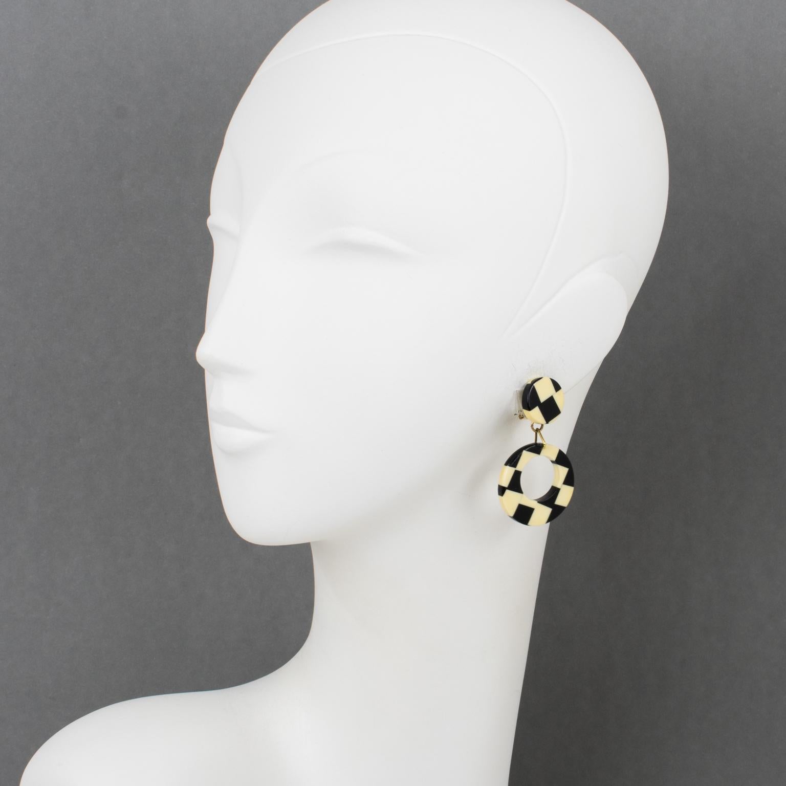 These stunning 1960s Pop Art clip-on earrings feature a dimensional dangling geometric donut design made of Galalith with a gorgeous checkerboard laminated pattern in black and off-white colors. These earrings have the specific French clip back used