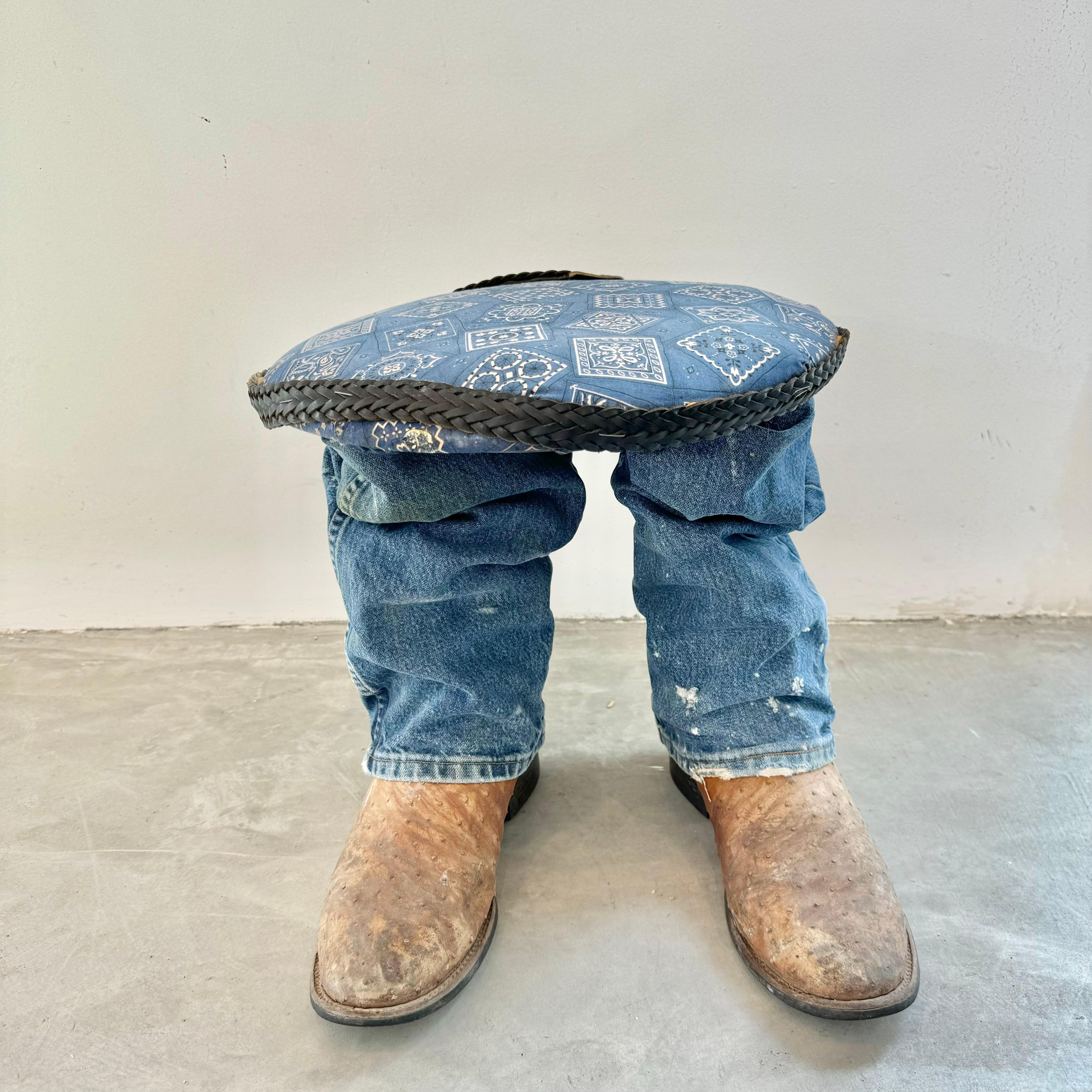 Unusual footstool with two legs covered with worn denim worn over a pair of rugged Ostrich leather cowboy boots as feet. Wooden circular top with a paisley blue bandana upholstery and a leather woven belt around the edge of the seat. Fun piece of