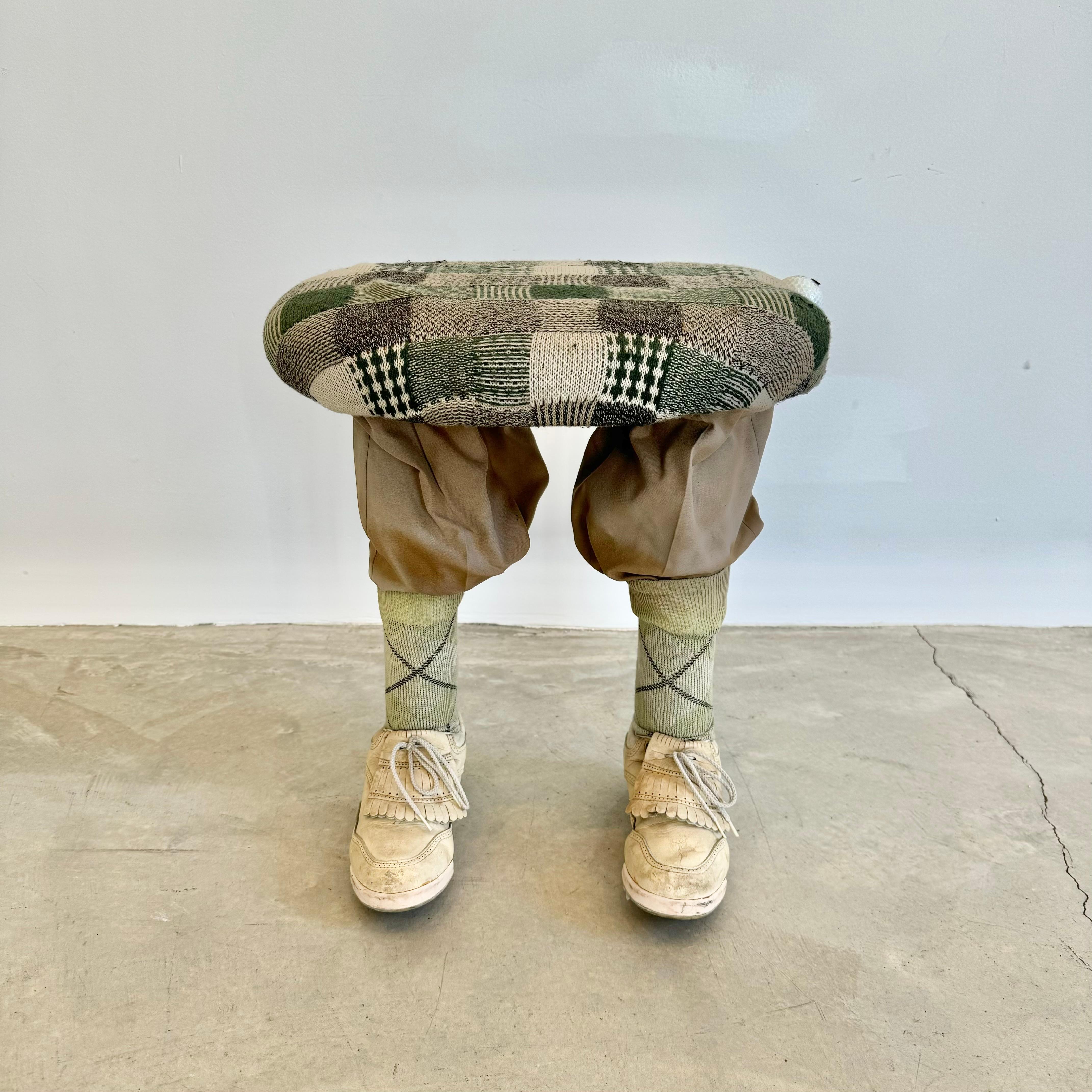 Insolito pouf con due gambe coperte da pantaloni kaki infilati in calzini alti con motivo Argyle e scarpe da golf in pelle color crema come piedi. Piano ovale in legno con rivestimento in tessuto patchwork. Palla da golf avvitata sul bordo.