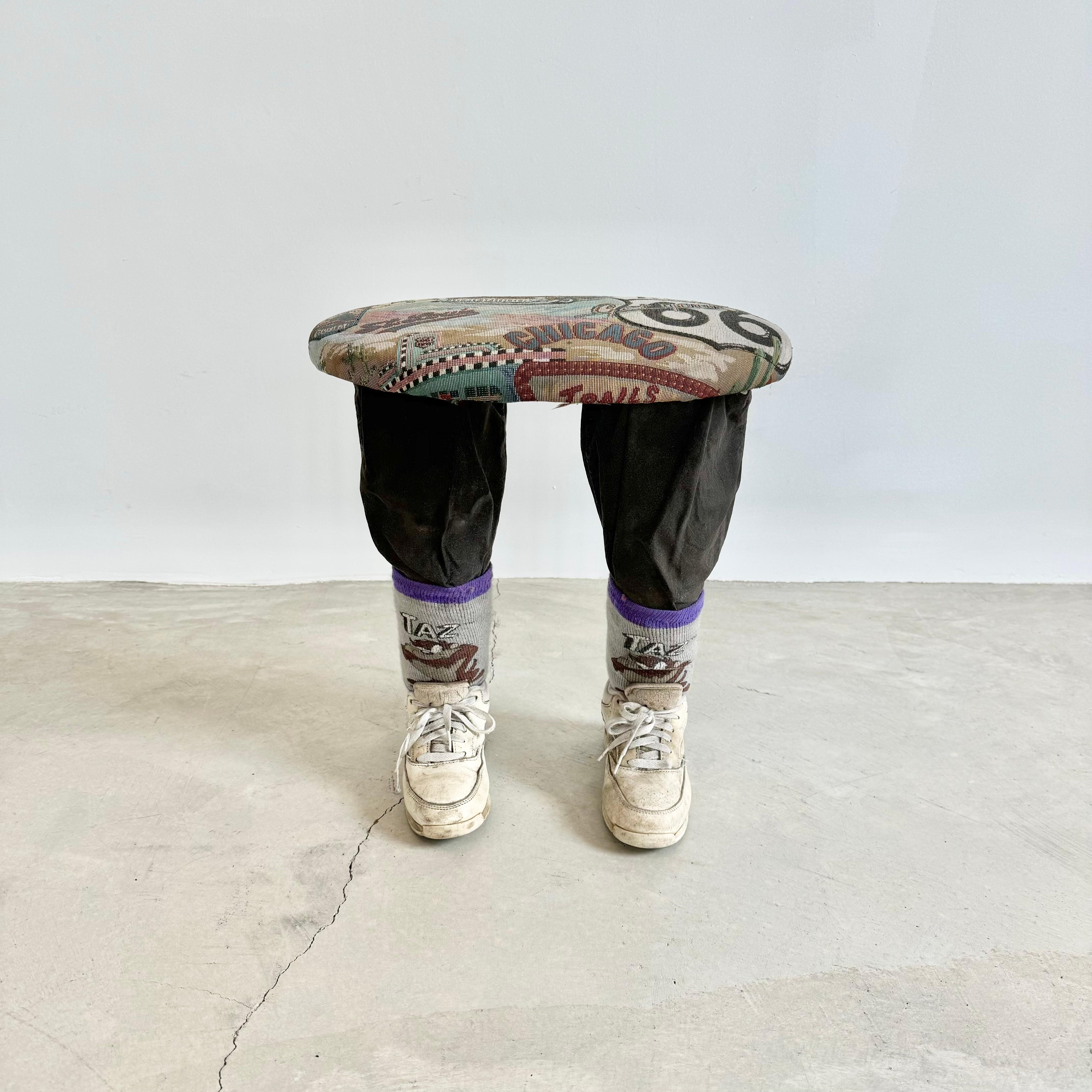 Unusual footstool with two legs covered with faded black denim, Taz cartoon socks and old LA Gear sneakers in white as feet. Wooden ovular top with a Route 66 fabric upholstery. Extremely sturdy and solid. Fun piece of pop-art and functional stool.