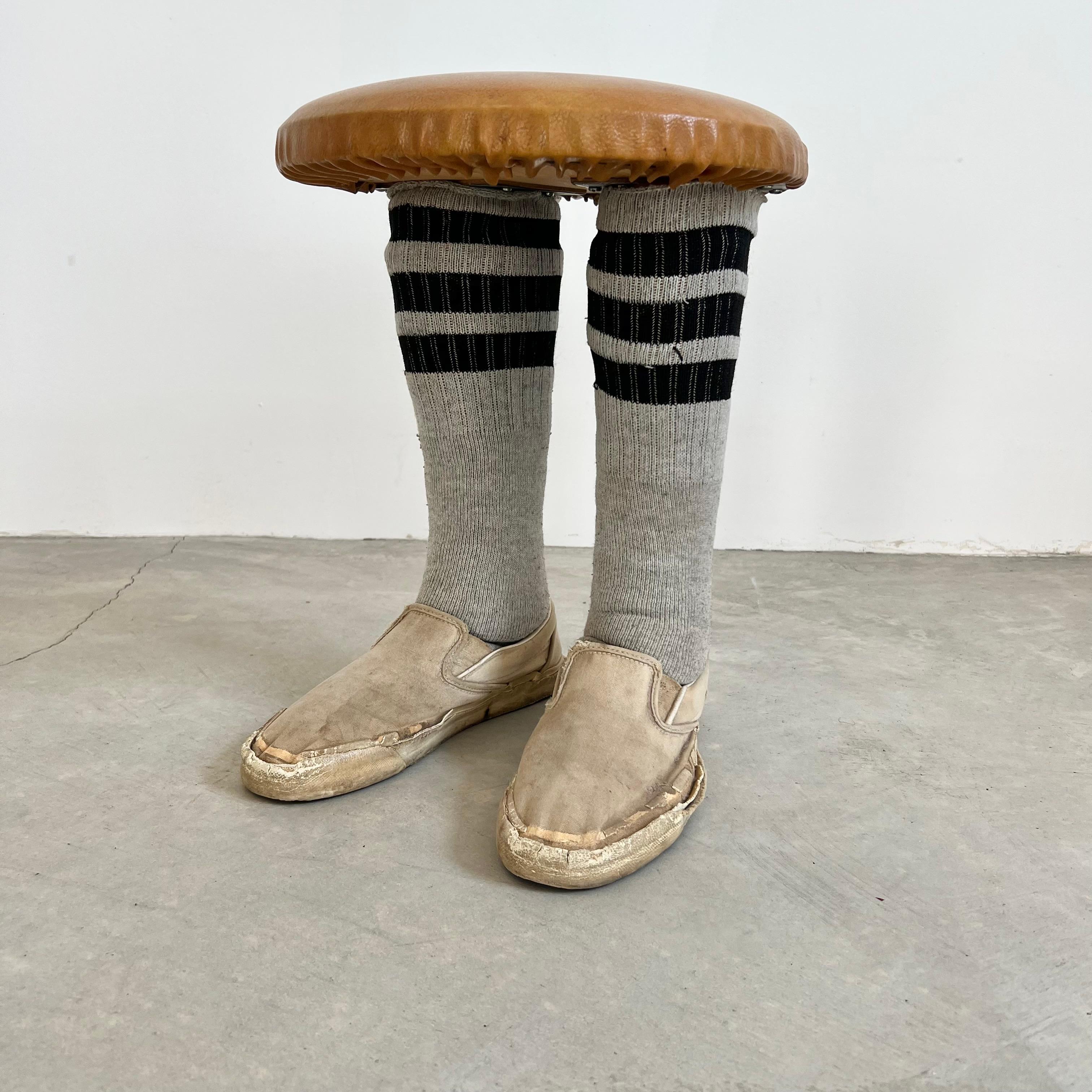 Unusual footstool with two legs wrapped in socks and old slip on sneakers as feet. Leather circular top. Extremely sturdy and solid. Fun piece of pop-art and functional stool. Good vintage condition with wear as shown.
 