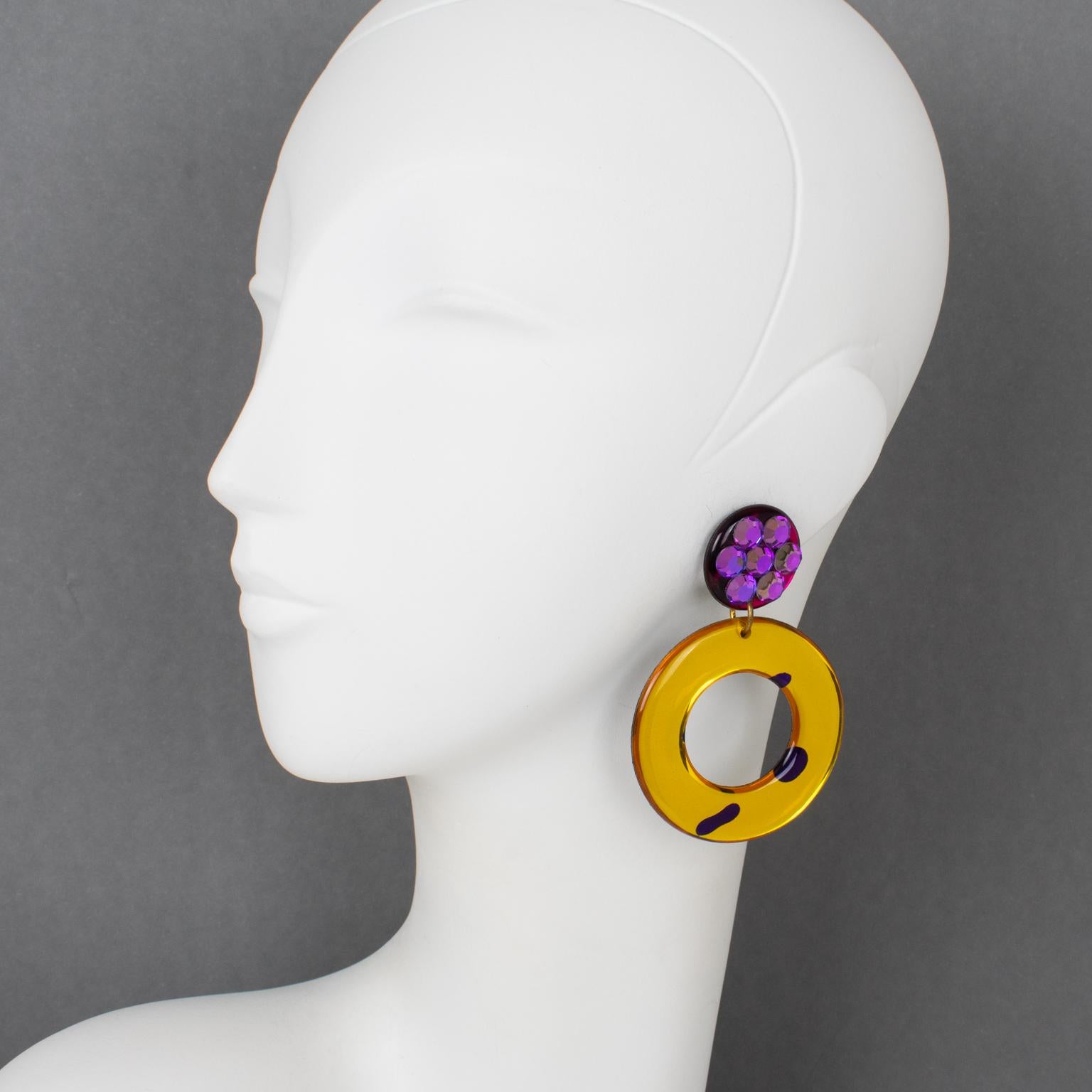 These stunning Lucite or Resin dangling clip-on earrings feature an oversized geometric design built with donut shapes in yellow saffron, amethyst purple, and hot fuchsia pink colors with a mirror effect and textured pattern. The earrings are ornate