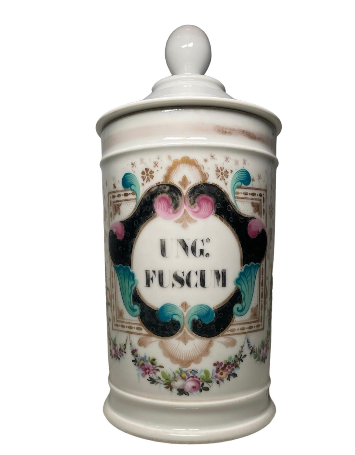 Questo è un vaso da farmacia in porcellana. Raffigura un vaso cilindrico di porcellana con coperchio a sfondo bianco e dipinto a mano nella parte anteriore con ampie volute nere che terminano con conchiglie turchesi e rosa che formano un cerchio che