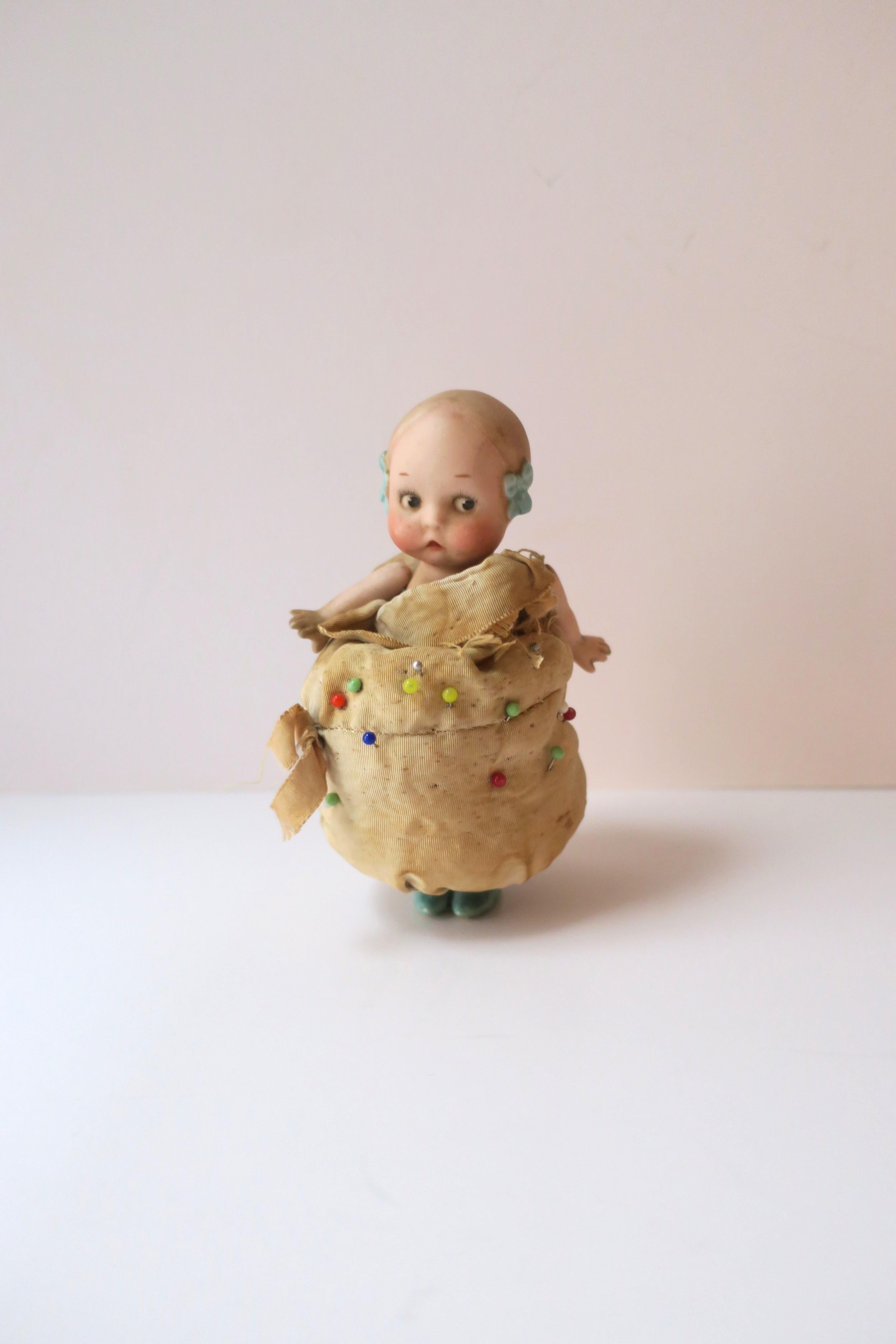 Coussin à épingles en porcelaine représentant une petite fille, vers le début du 20e siècle. Cette poupée en porcelaine, avec une robe en coussin d'épingles, est représentée avec un beau visage, des joues roses, des nœuds bleus dans les cheveux, un