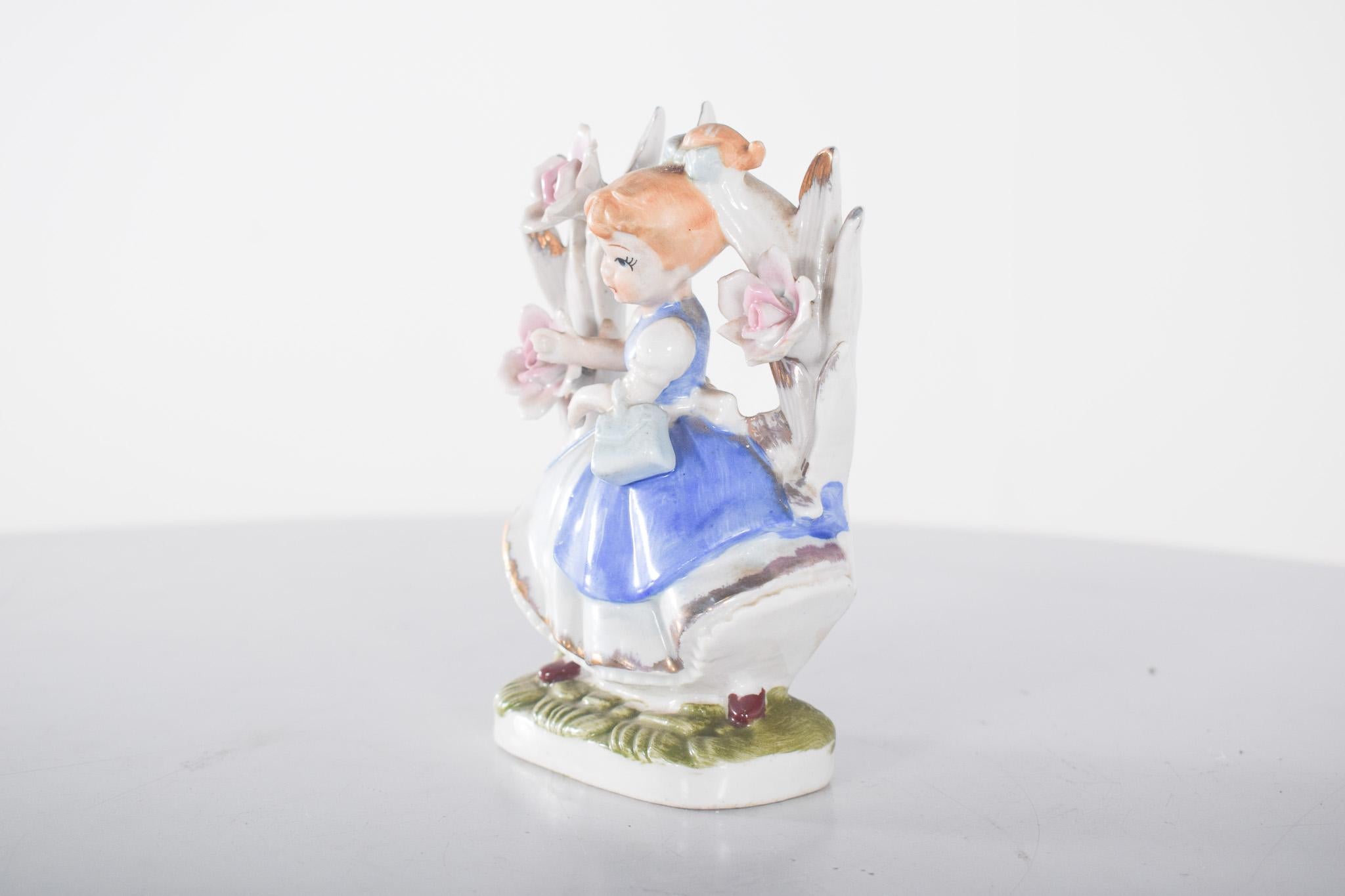 Figura de porcelana de una niña sentada. Esta escultura representa a una joven con un elegante vestido azul y blanco decorado con oro. Lleva el pelo rubio recogido con un lazo azul claro y su expresión facial transmite suavidad e inocencia. Sostiene