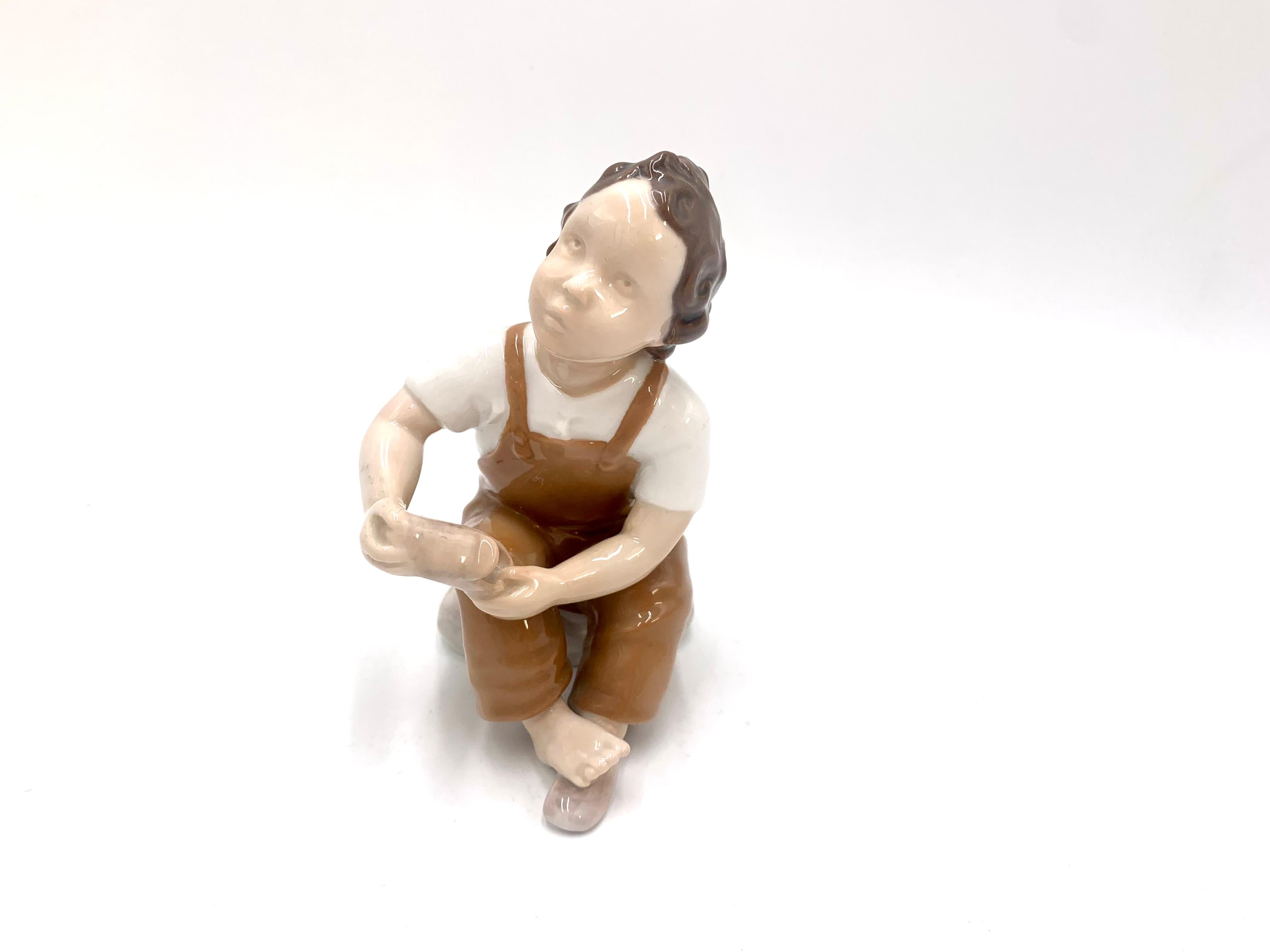 Figurine en porcelaine d'un garçon demandant de l'aide pour mettre sa chaussure

Fabriqué au Danemark par Bing & Grondahl

Produit en 1958-1962

Numéro de modèle : 2275

Très bon état, aucun dommage

Mesures : hauteur 13,5 cm, largeur 7
