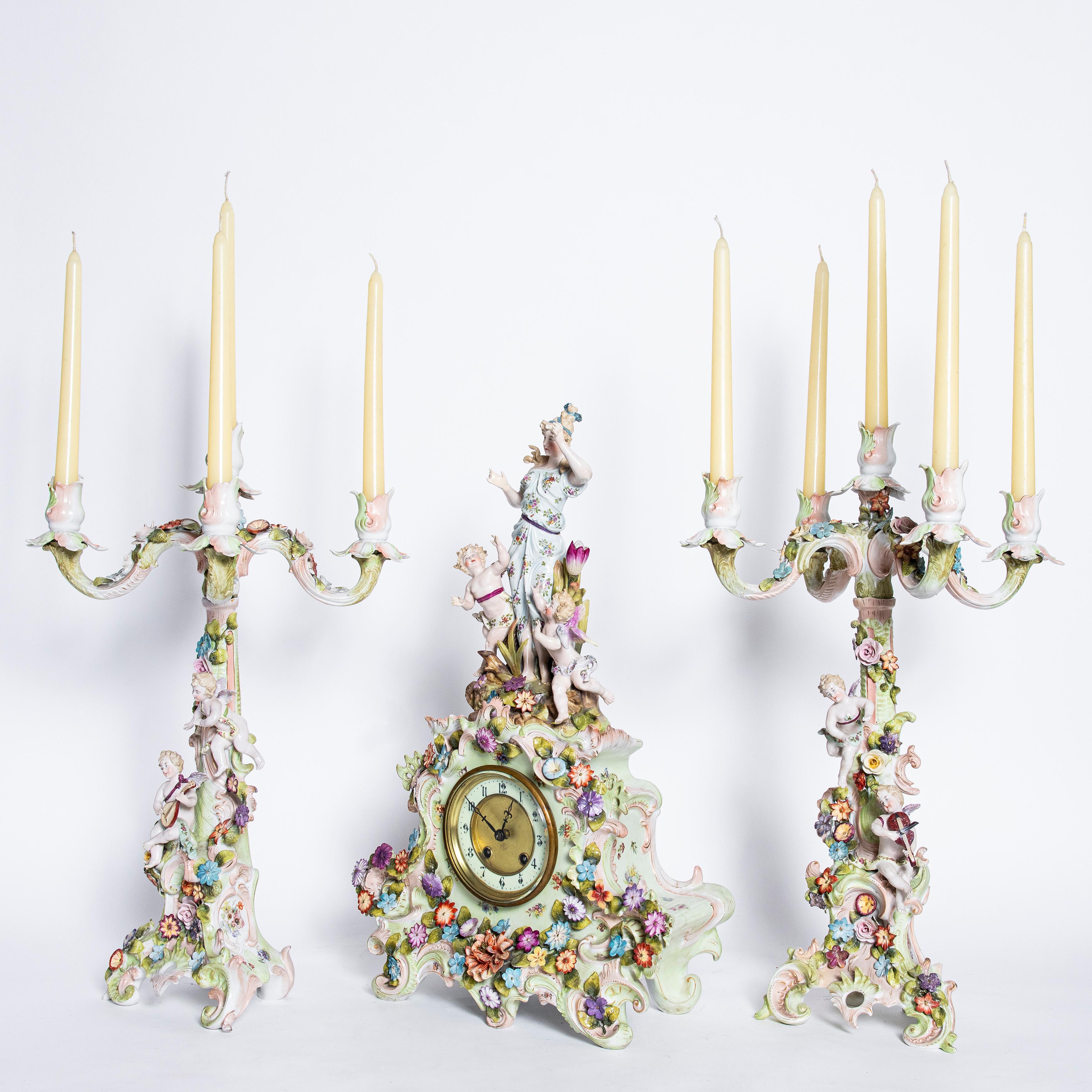 Porcelain garniture with flowers and angels. Germany, circa 1900.

Candelabras dimensions: 26 cm depth, 27 cm width, 52 cm height.
Clock dimensions: 16 cm depth, 36 cm width, 54 cm height.