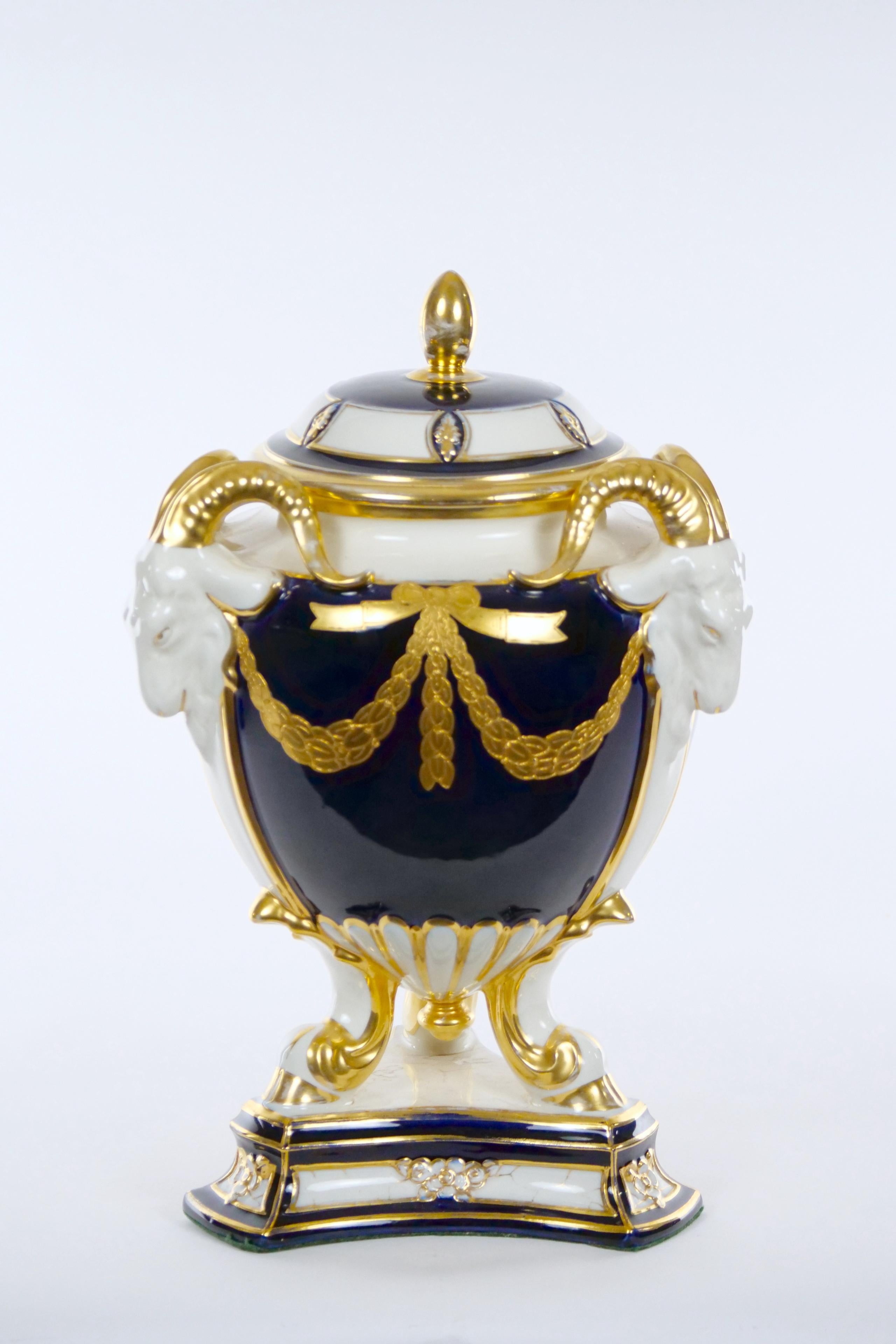Stunning glazed porcelain / hand painted and gilt decorated covered urns with figural Ram’s head handles. The urn features a deep glazed navy blue with an egg shell background decorated with gilt gold resting on a geometric shape form base. The urn