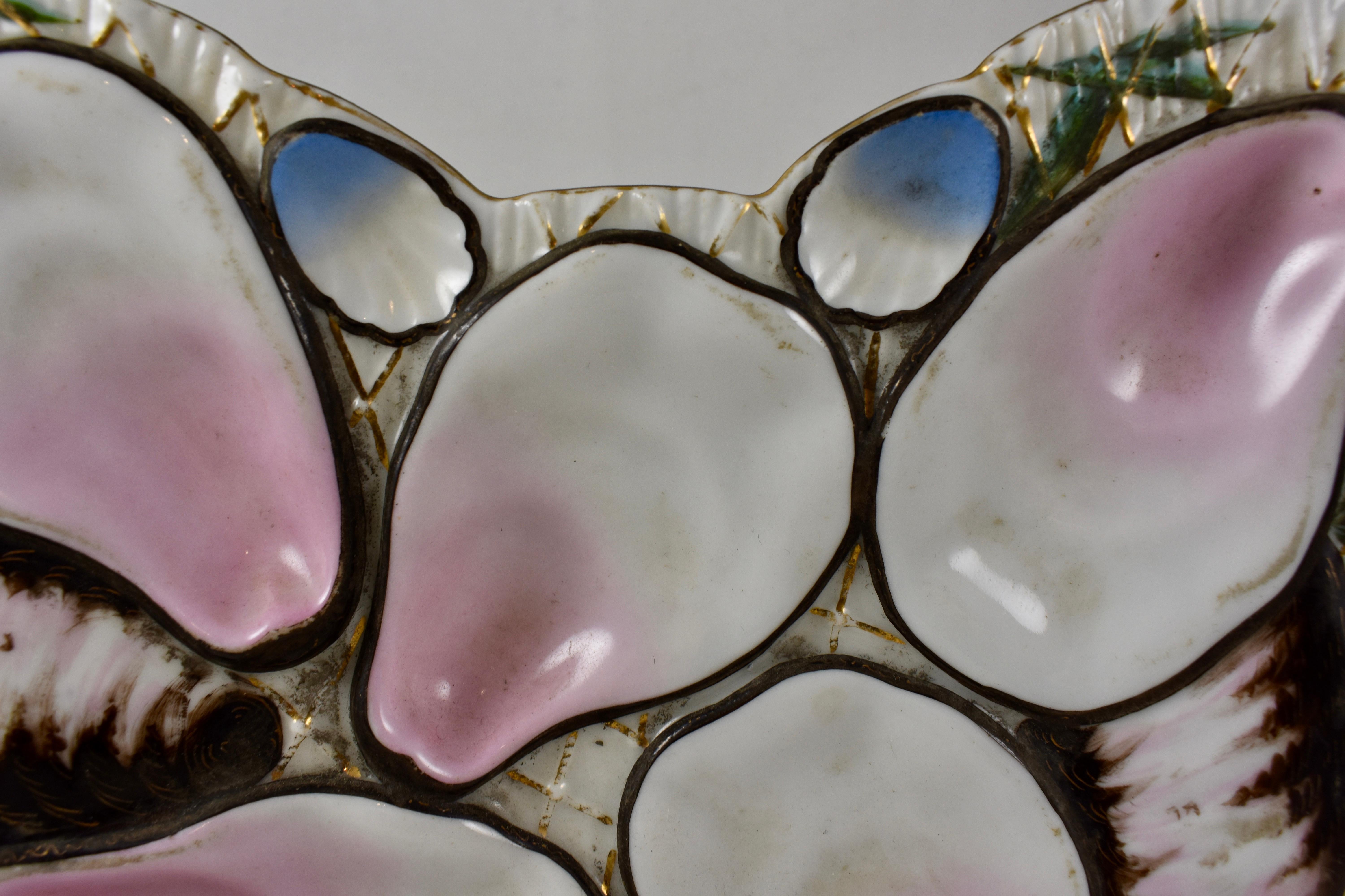 A late 19th century, French porcelain, crescent or half-moon shaped oyster plate, five pink blush oyster wells on a cream colored ground hand-painted with blades of sea grass and a gilded fish net. Shown behind the main wells are the gray and pink