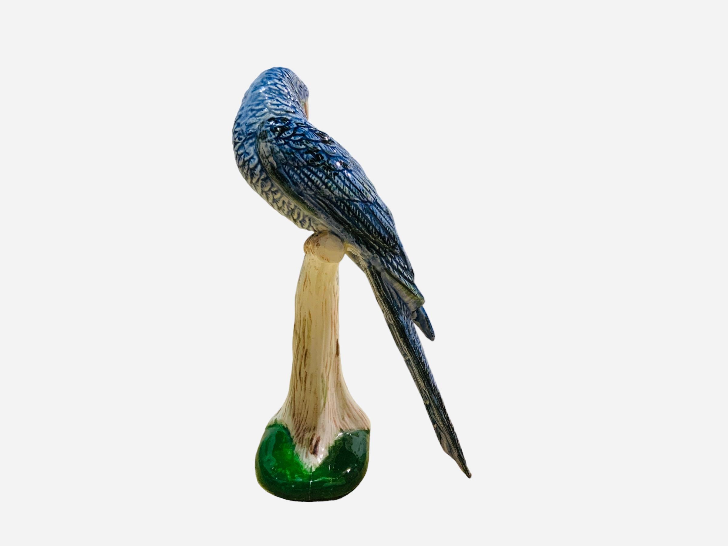 This is an unmarked porcelain figurine of a bird. It depicts a very well done hand painted blue parrot . It is standing up in a branch of a trunk supported by a green oval base.