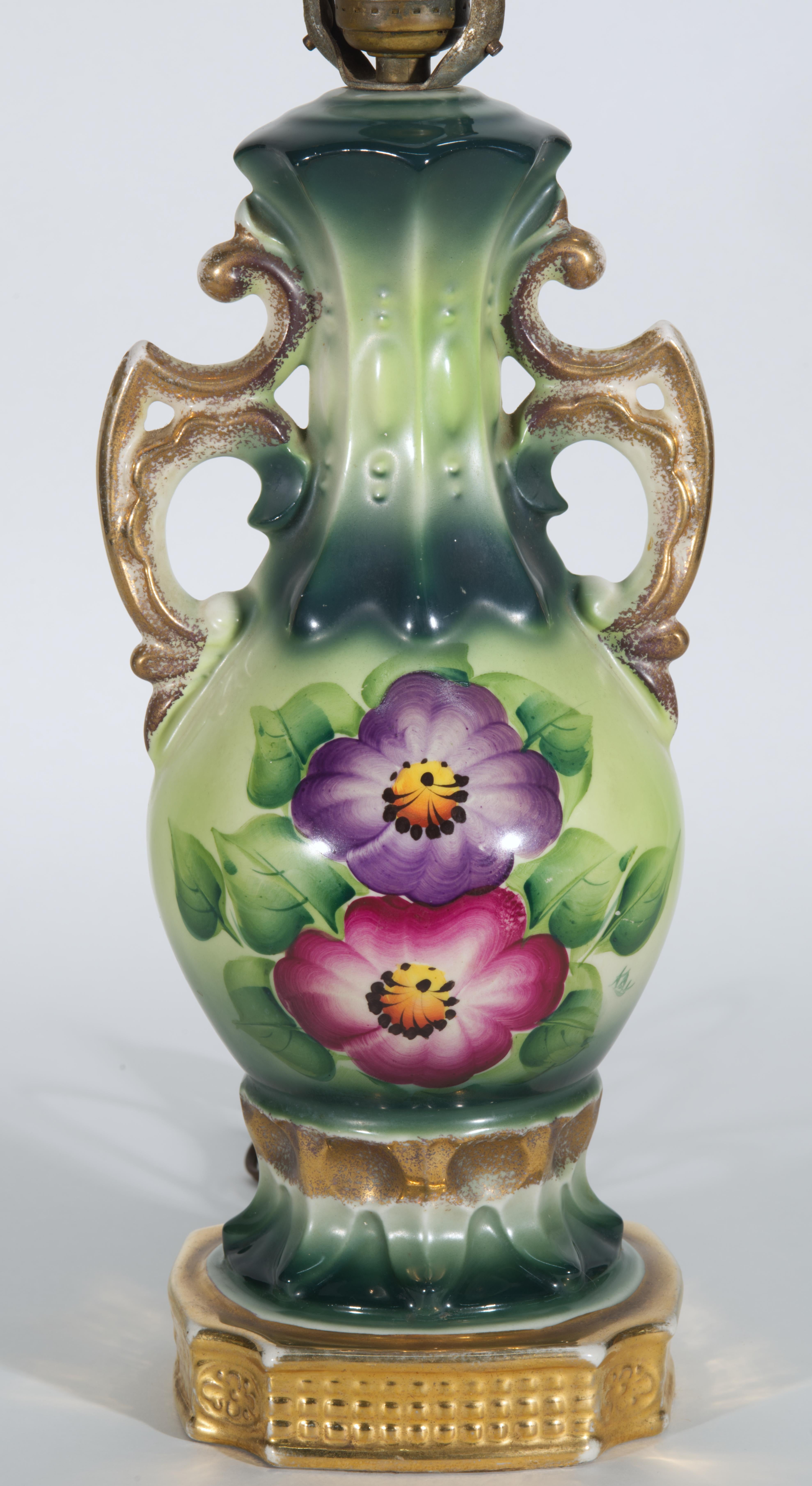 Striking large antique or vintage table lamp features urn shaped porcelain body with two ornate decorative twin handles, hand-painted in Victorian style with large flowers, surrounded by leaves. Prominent pink and purple flowers with yellow and