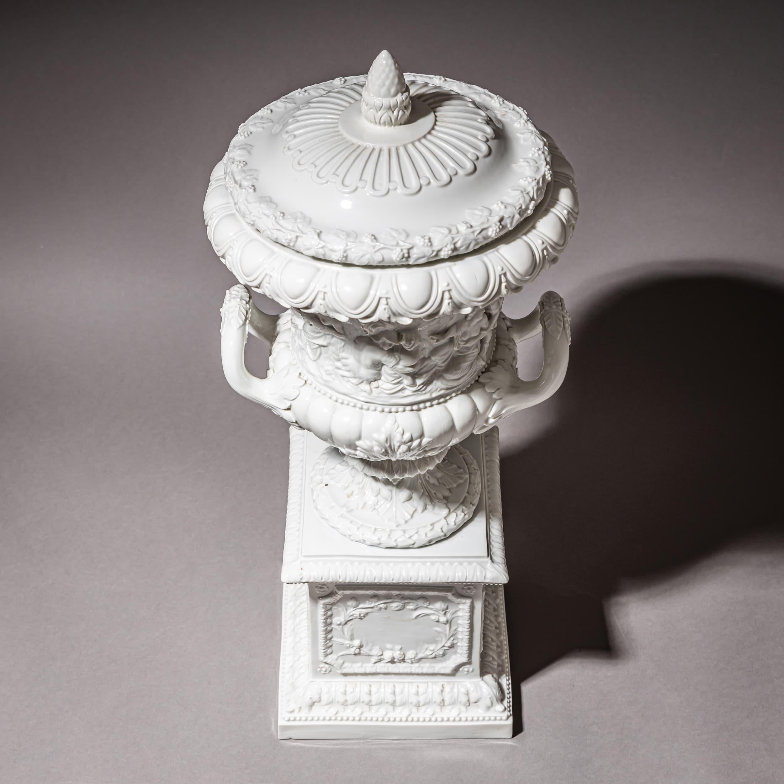 Large lidded urn of white-glazed porcelain on a rectangular plinth with beaded molding and acanthus leaves. The body features a rounded cartouche with rosettes and a garland of roses in relief. Above rises a round foot with leaf decoration, leading