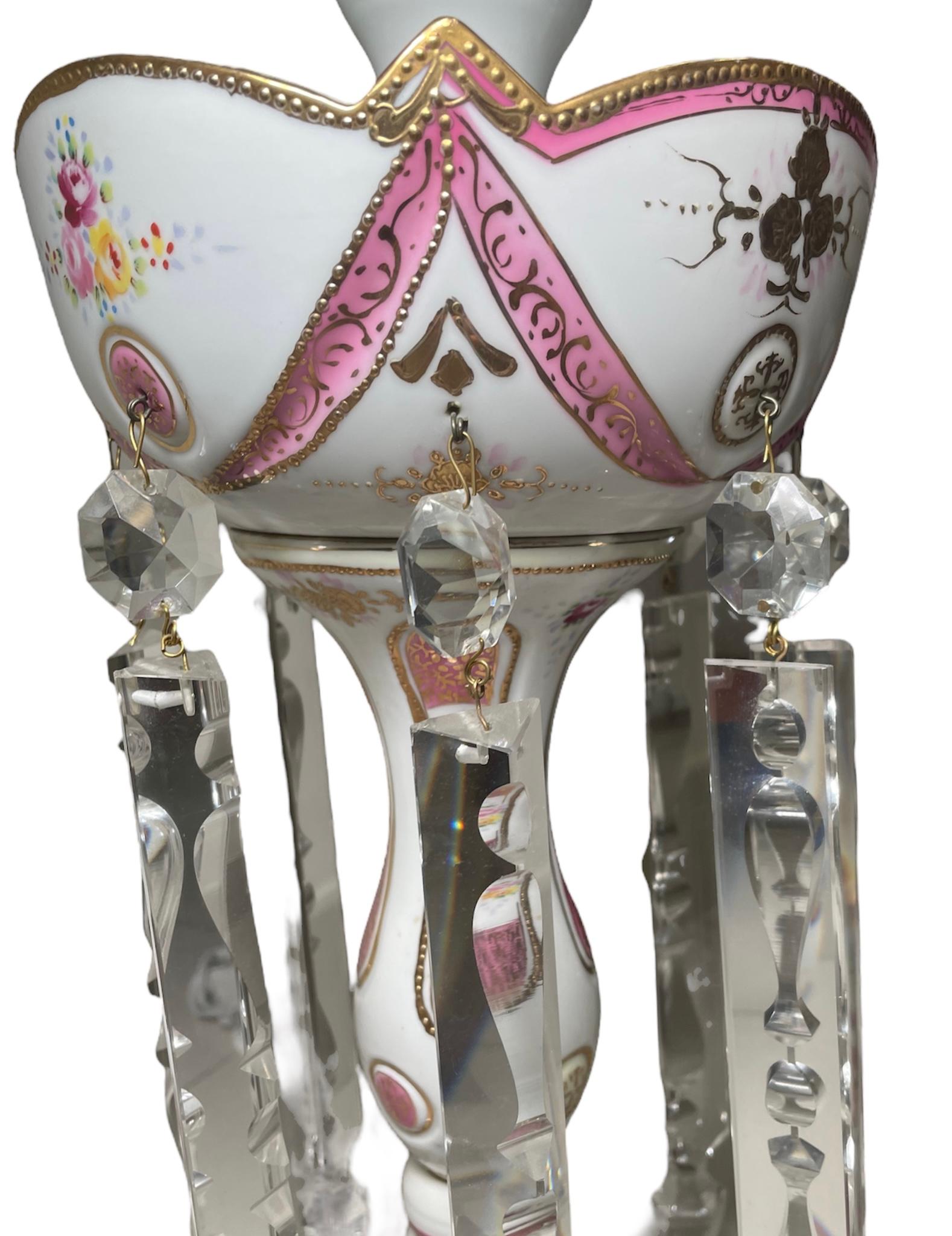 Dies ist eine Porzellan Mantle Luster Lampe. Es zeigt eine handgemalte Lampe mit weißem Hintergrund, die durch rosa Bänder, Girlanden und Kreise mit vergoldeten Details verziert ist. Ein Rosenstrauß schmückt auch das Dekor der Lampe. Der obere Rand