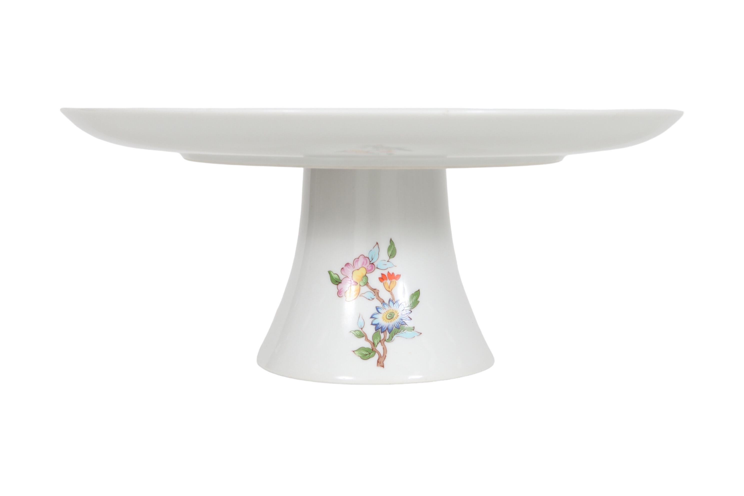 A white porcelain pedestal cake stand in the Le Jardin pattern, made by Dolphin Fine China of Japan. Decorated with delicately painted and brightly colored pink, blue and orange flowers amid intertwining foliage on the plate, and with bouquets of