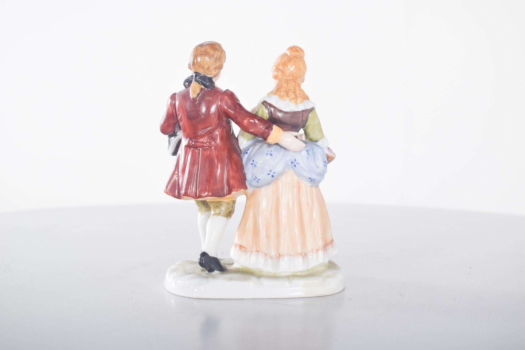 Scultura in porcellana raffigurante una signora e un gentiluomo. Decorazione policroma dai toni forti. La figura femminile indossa un abito lungo nei toni tenui del blu e del beige, con un corpetto marrone aderente e mezze maniche. Tiene un cesto di