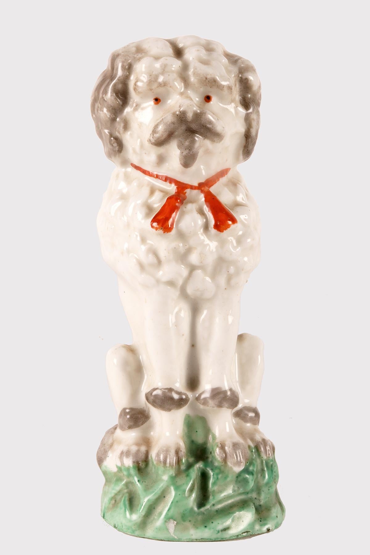 Bemalte Porzellanskulptur, die einen im Gras sitzenden Pudelhund mit einer roten Schleife um den Hals darstellt. England um 1900.
