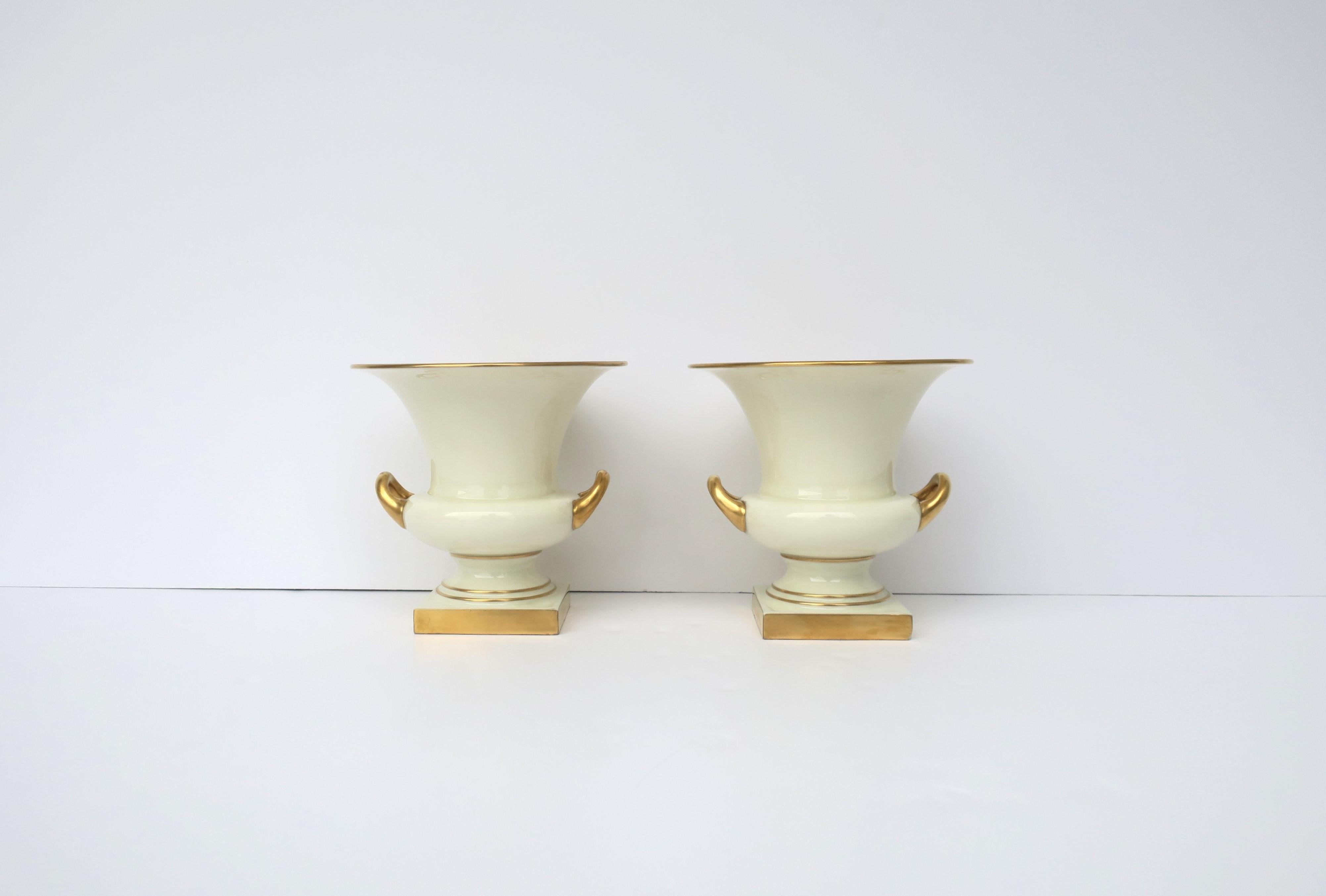 A pair of cream and gold porcelain urns in the Neoclassical design style, circa mid to late-20th century, USA. Urns are a cream porcelain with gold detail on handles and bases. Urns can also double as vases. A great set for any table, fireplace