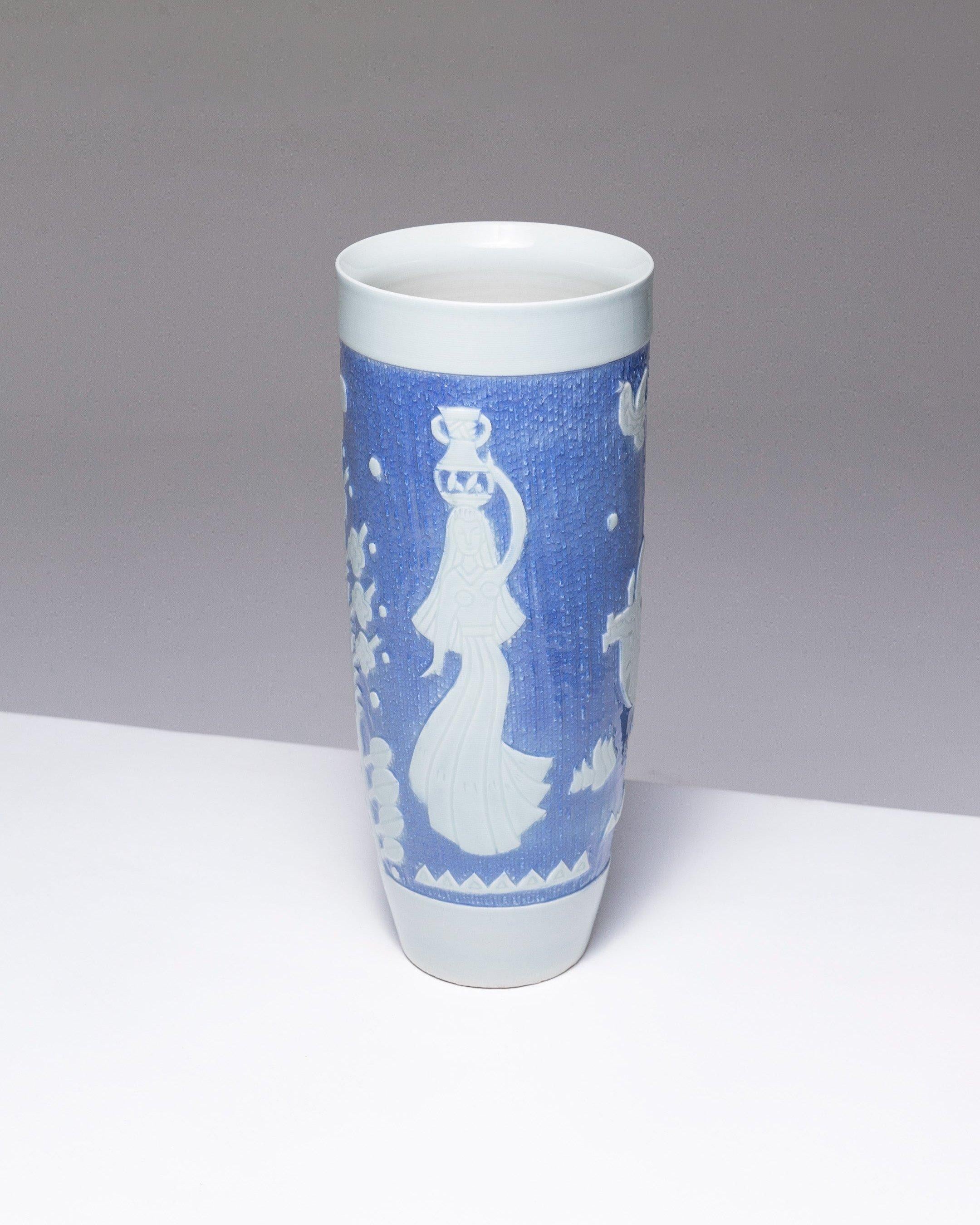Vaso in porcellana con corpo circolare svasato verso l'alto, senza collo. Anni '50, Nord Europa. Su uno sfondo blu leggermente sgranato, una decorazione stilizzata rotante in leggero rilievo bianco: Una giovane donna che porta un vaso e alcuni