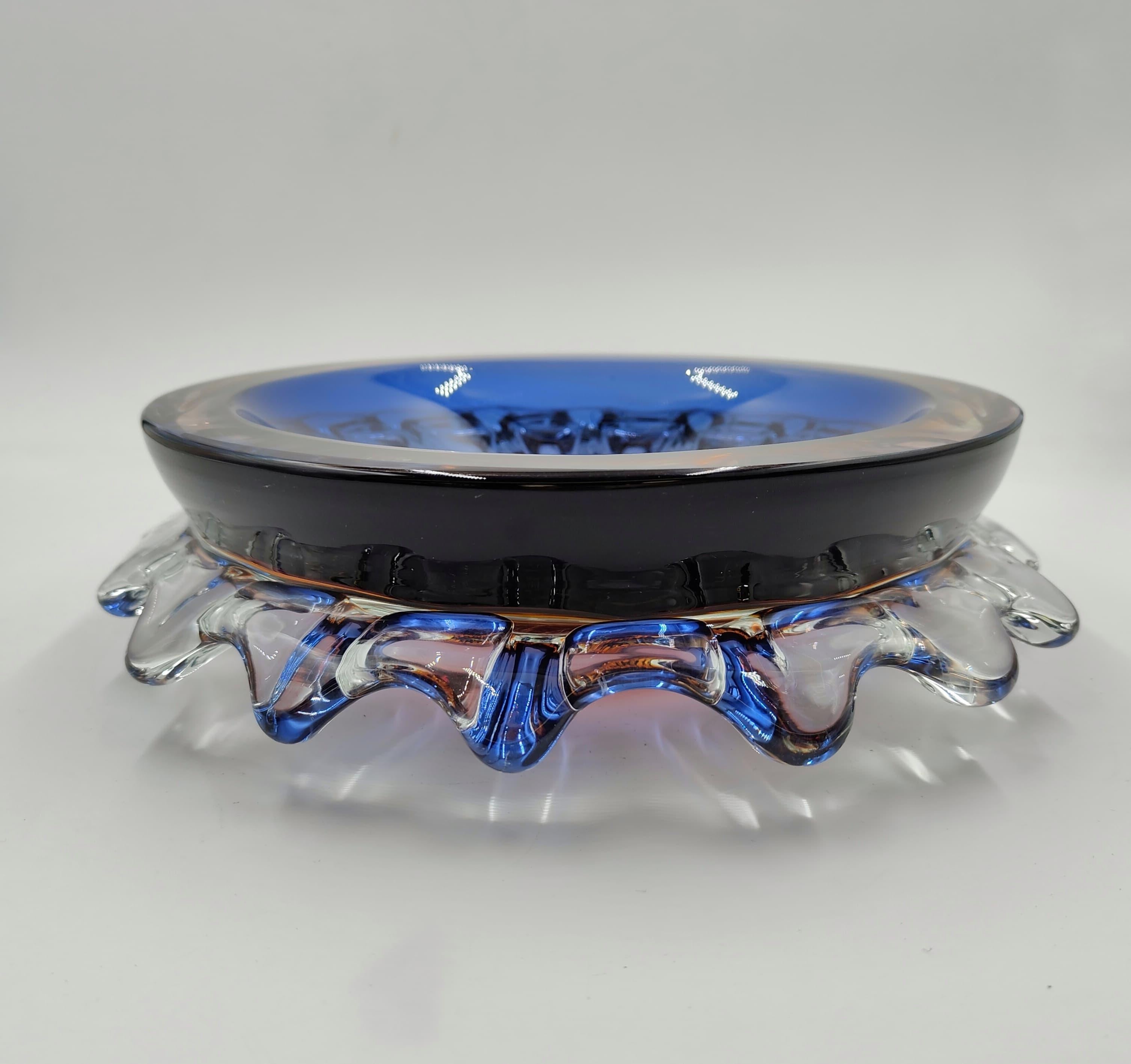 Dieser Halter ist aus Muranoglas aus den 1980er Jahren gefertigt. Das von Hand modellierte Werk bringt die Handwerkskunst der Glasmacher aus Murano zur Geltung und kombiniert Formen und Farbtöne.
Der zentrale Teil des Objekts hat eine Färbung, die