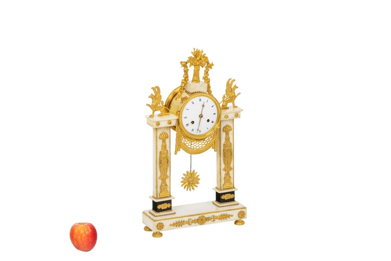 Portico clock in chiseled and gilt bronze composed of a rectangular base in white marble. Architecture with two columns topped by griffins holding the dial. The columns are decorated with two Egyptian goddesses and foliage. Dial in white enamel
