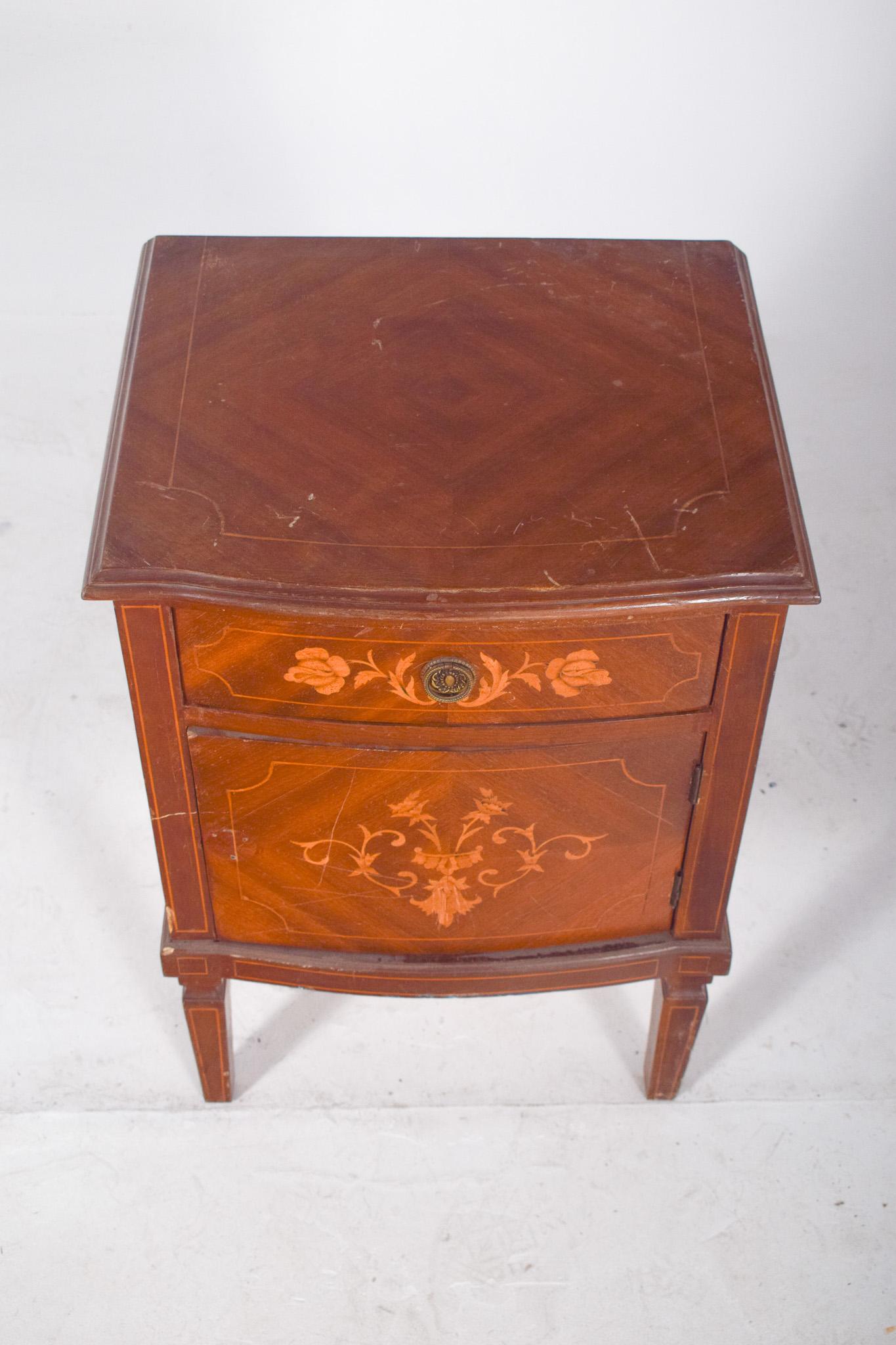 Bedside table with marquetry work. It consists of an upper drawer with an ornate round metal handle, decorated with carved flowers in lighter tones, and a door with a marquetry front centred on a symmetrical floral pattern, framed by fine fillets.