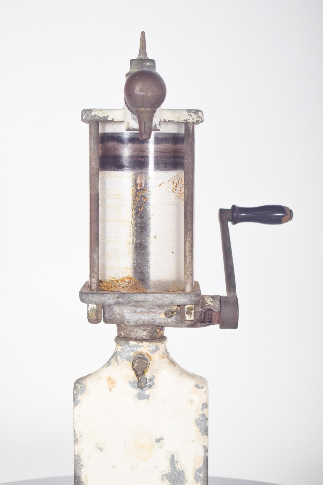 Distributeur manuel d'huile d'olive. Il s'agit d'un équipement ancien, principalement en métal, dont la structure est robuste et qui présente des signes d'usure au fil du temps. Il possède un réservoir cylindrique transparent où l'huile est stockée,