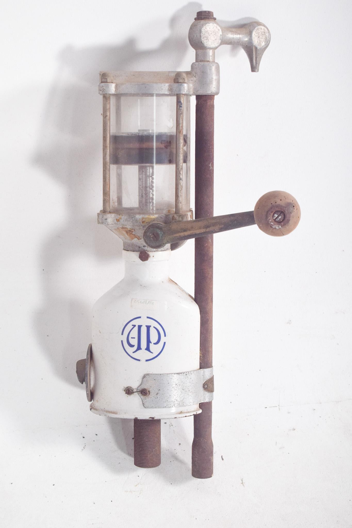 Distributeur manuel d'huile d'olive. Il s'agit d'un équipement ancien, principalement en métal, dont la structure est robuste et qui présente des signes d'usure au fil du temps. Il possède un réservoir cylindrique transparent où l'huile est stockée,
