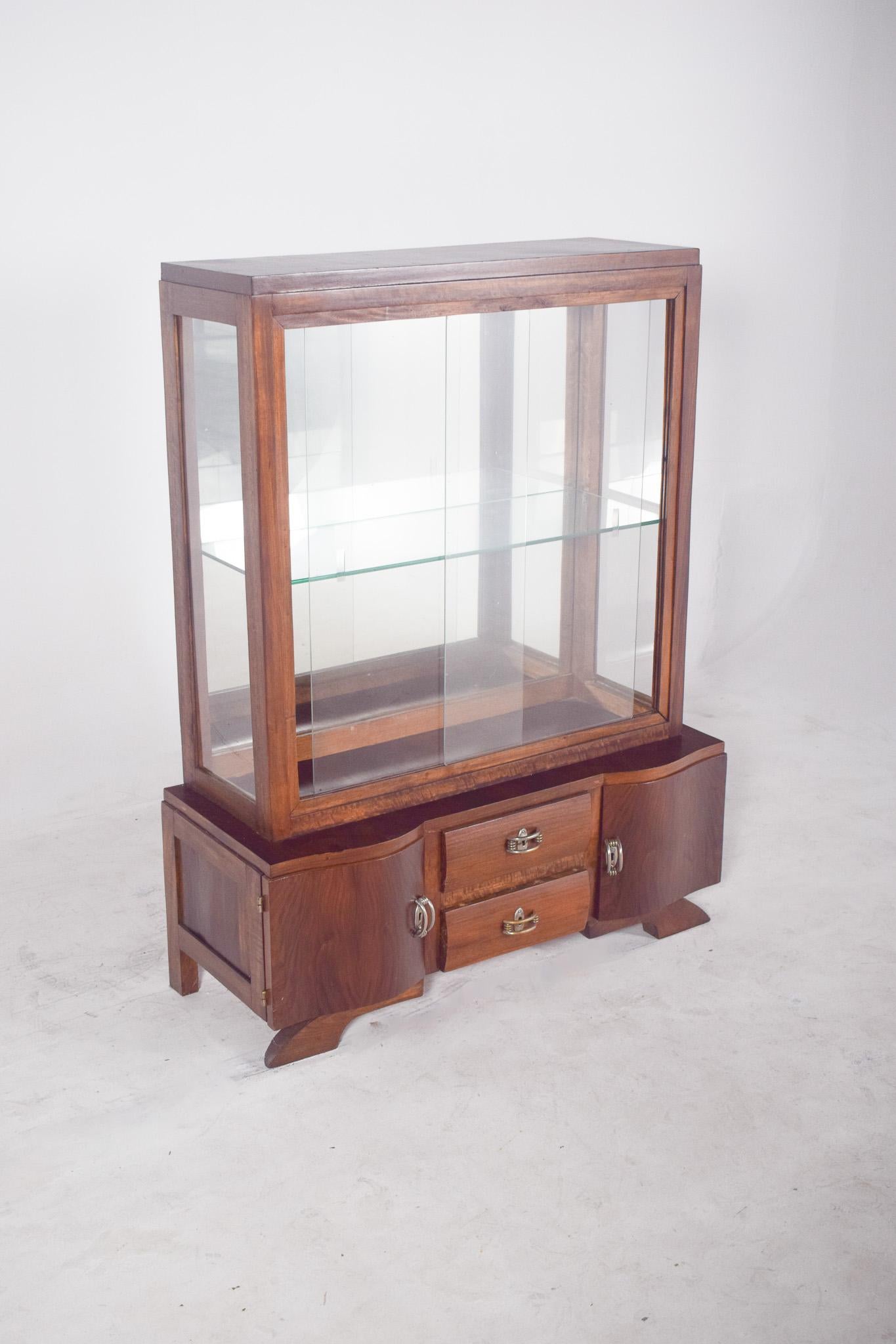 Art Deco-Vitrine. Das Oberteil besteht aus einem verglasten Fach mit einem Glasboden und Schiebetüren, ideal für die Präsentation von Dekorationsstücken, Sammlerstücken oder feinem Porzellan.
Die Basis des Schranks besteht aus zwei funktionalen