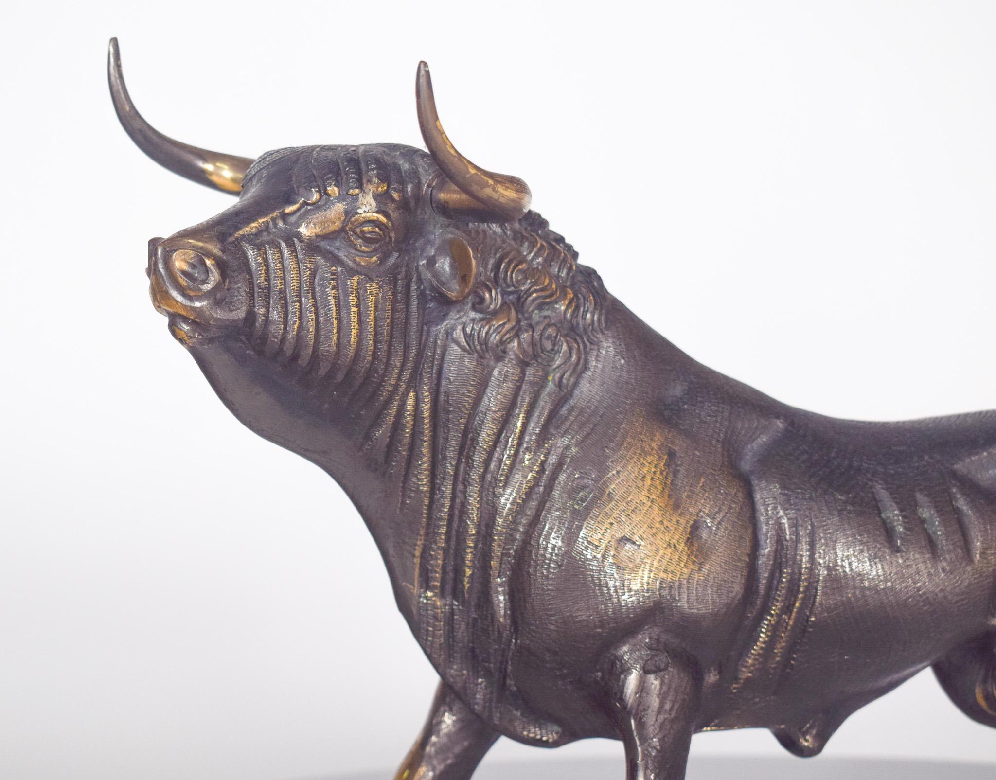 Brass bull sculpture. With an aged finish that combines dark tones and golden brass details. The sculpture depicts a bull in an attacking position, with its head slightly raised, nostrils open and muscles well defined, conveying a sense of strength