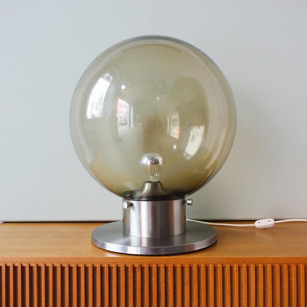 Cette lampe de table de style brutaliste a été conçue par Francisco Conceição Silva et produite au Portugal, dans les années 1970. La base est en métal chromé brossé, où un grand verre soufflé à la main est placé. Elle peut être utilisée comme lampe