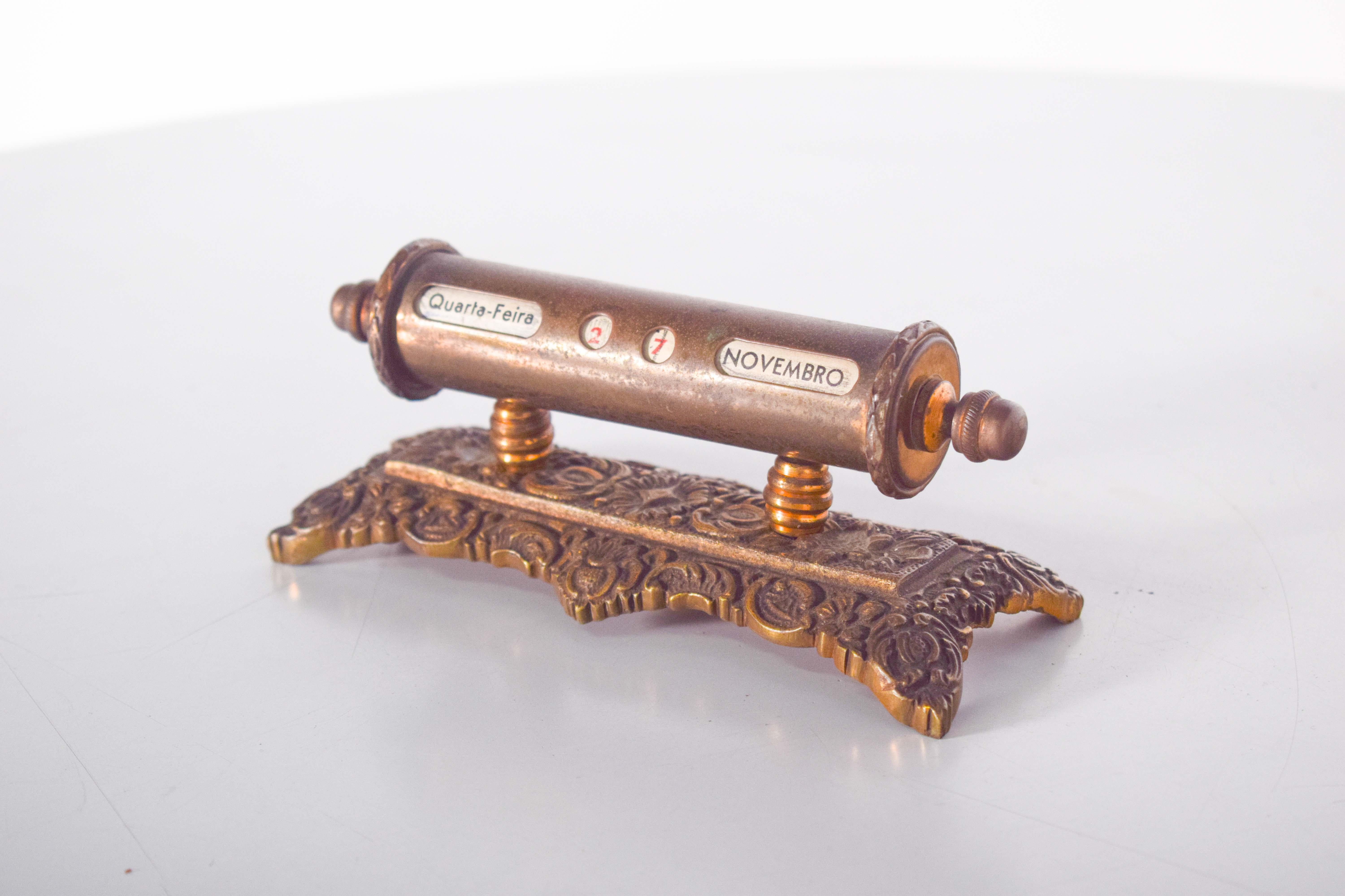 An elegant vintage desk calendar, designed in the form of a roll, elegantly supported by a golden metal base. The calendar features beautifully embossed floral patterns, adding a touch of charm and sophistication. It displays the day of the week,