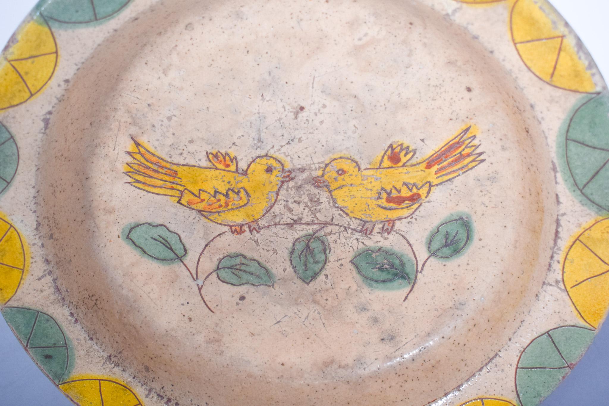 Plaque d'argile, peinte et émaillée. Décor polychrome avec deux oiseaux, peint à la main, caractéristique de la tradition artisanale. La surface présente un fond beige clair, avec un design central de deux oiseaux jaunes perchés sur des branches