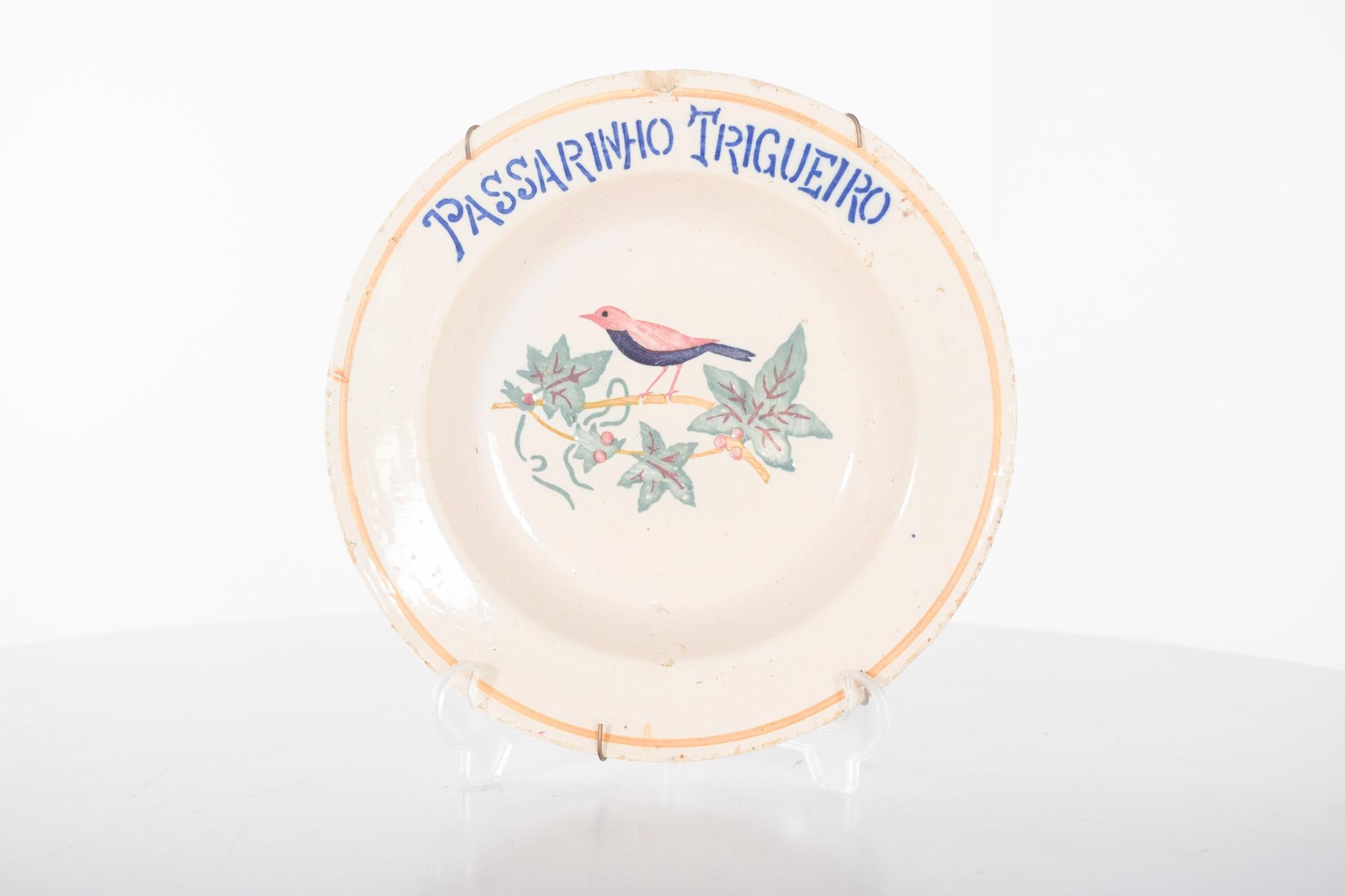 Plat en faïence portugaise avec oiseau. Décor polychrome dans des tons doux avec un petit oiseau perché sur une branche avec des feuilles et des baies au centre. Sur le bord supérieur, l'inscription 