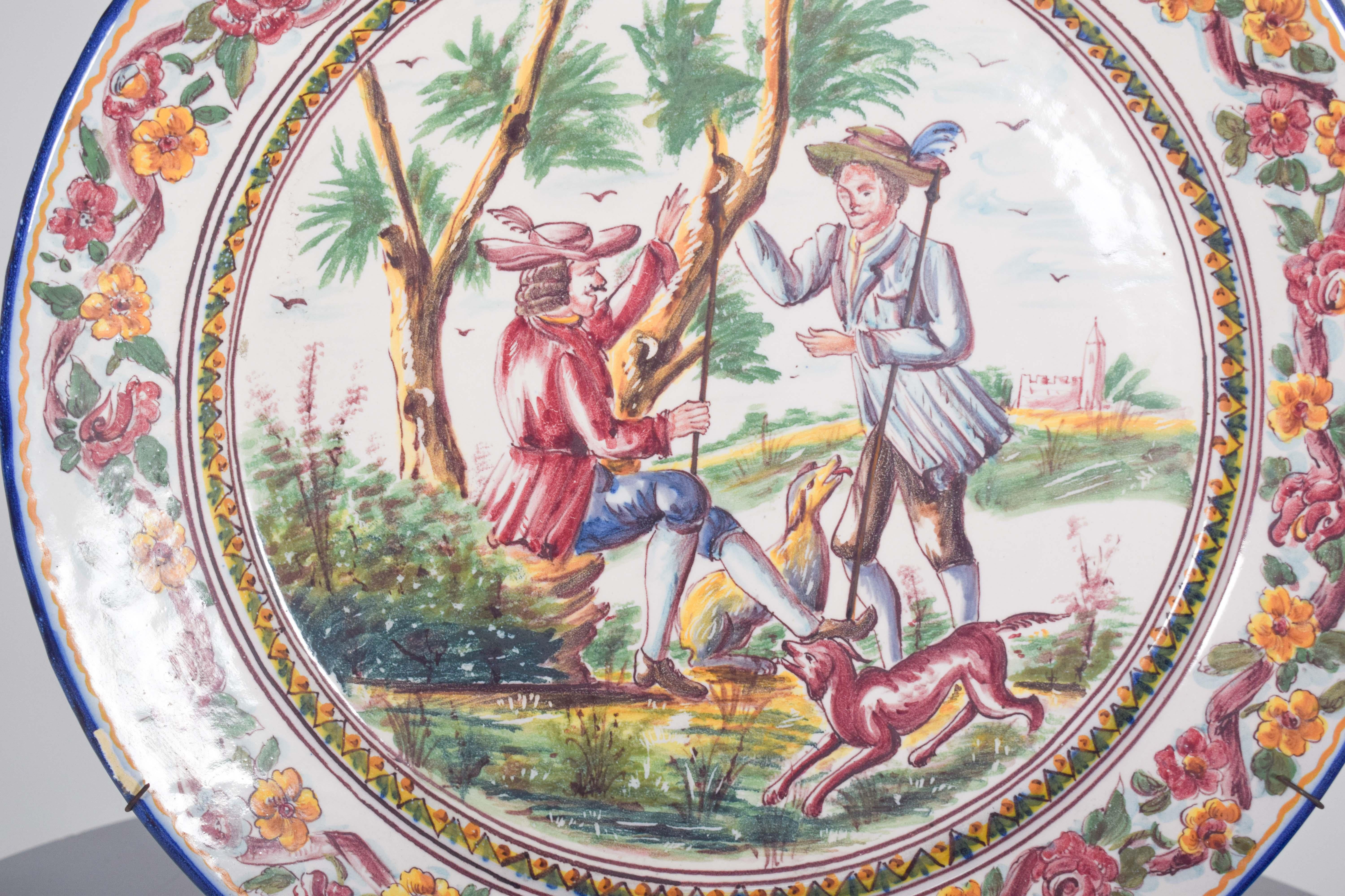Grande assiette, en faïence portugaise de Sant'Anna. Décor polychrome avec paysage et personnages. Au centre se trouve une scène rurale avec deux hommes portant des vêtements typiques du XIXe siècle, entourés d'un paysage d'arbres et de végétation.