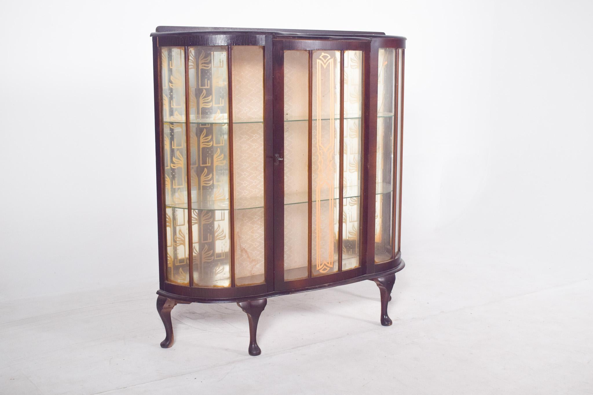 Vitrine avec pieds galbés. Il se distingue par sa forme incurvée et ses élégantes portes en verre qui révèlent un intérieur soigneusement conçu. La structure en bois foncé contraste harmonieusement avec le fond recouvert d'un tissu clair à motifs