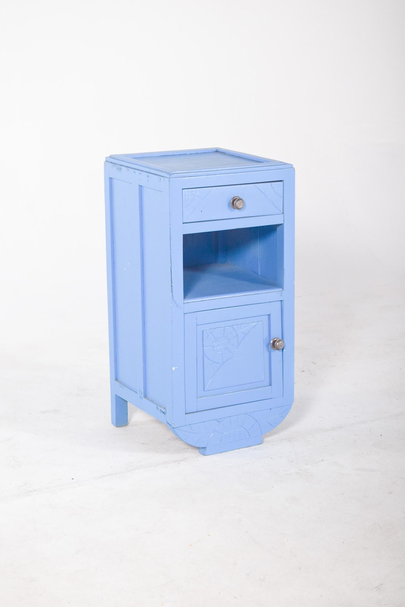 Bedside table painted light blue. With an upper drawer with a round metal handle, an open niche in the centre and a lower door with a similar handle. Carved details in relief with floral and geometric motifs on the corners of the drawer and door
