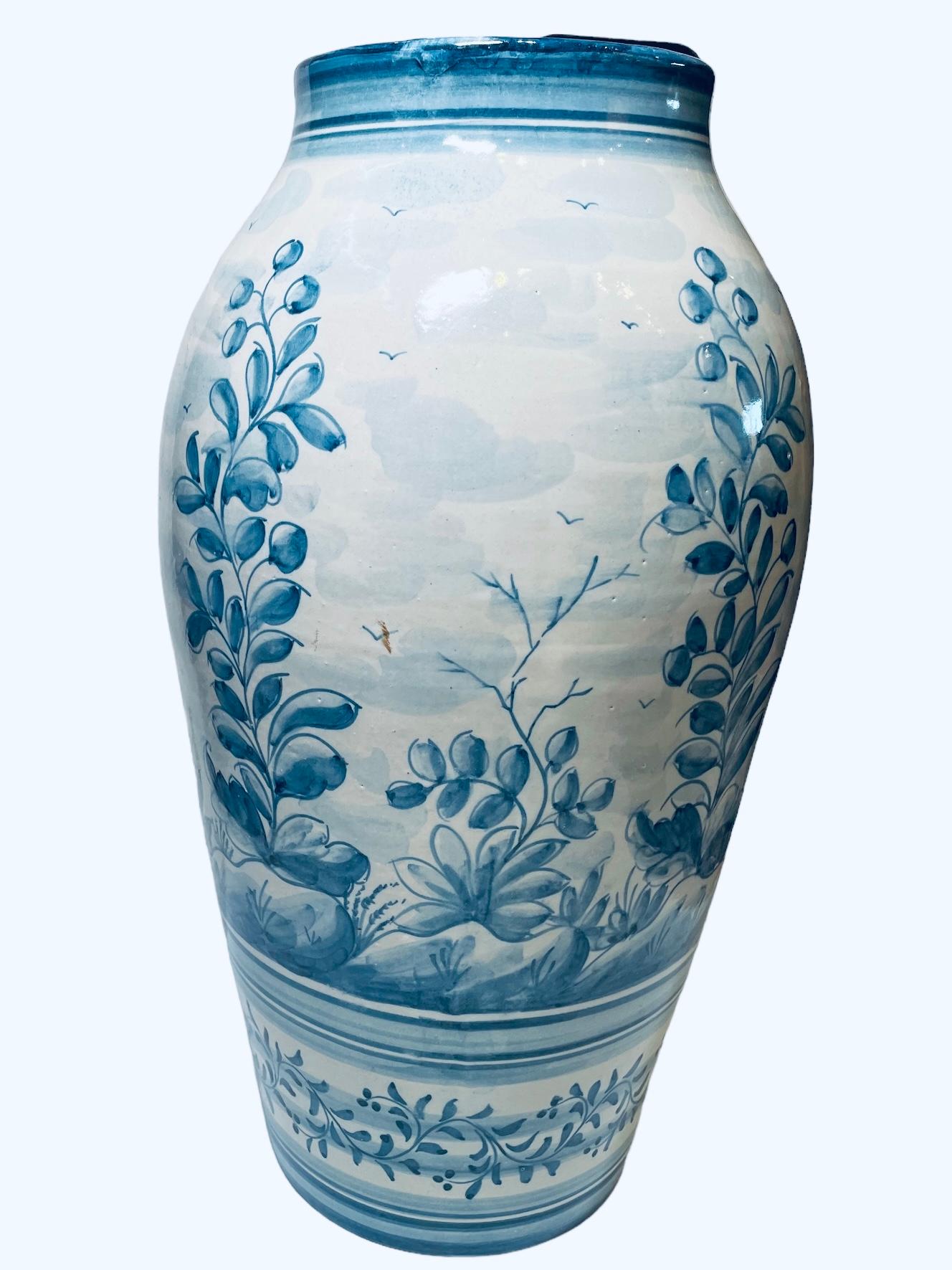 This is a Portuguese Majolica Vase Urn. It depicts a hand painted blue and white scenes of young olive trees and some birds flying in the sky. The bottom of the vase is adorned with a hand painted garland of a climbing plant. Below the vase is