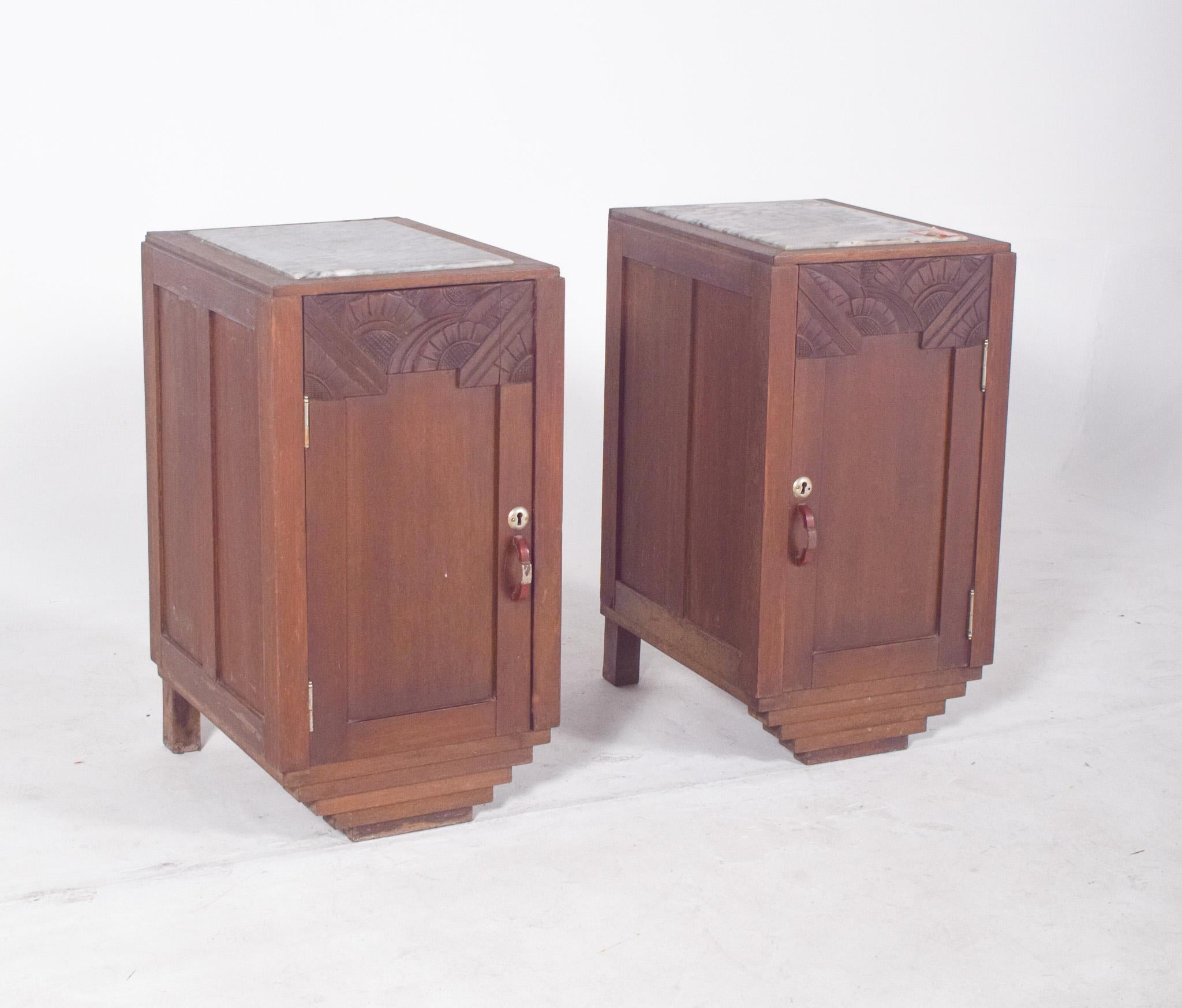 Pair of Deco-style bedside tables. They feature a front door with a simple wooden handle and metal lock, under a carved top panel with a geometric pattern in overlapping semicircles, typical of the decorative aesthetic of the 1930s. The stepped base