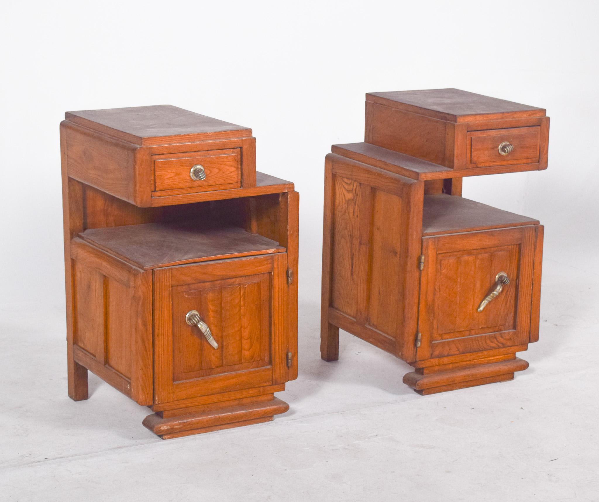 Pair of Deco-style bedside tables. They have raised tops that house small drawers with round metal handles, followed by open central niches and lower doors with ornate conical-shaped handles. The sides feature simple, discreet mouldings, while the