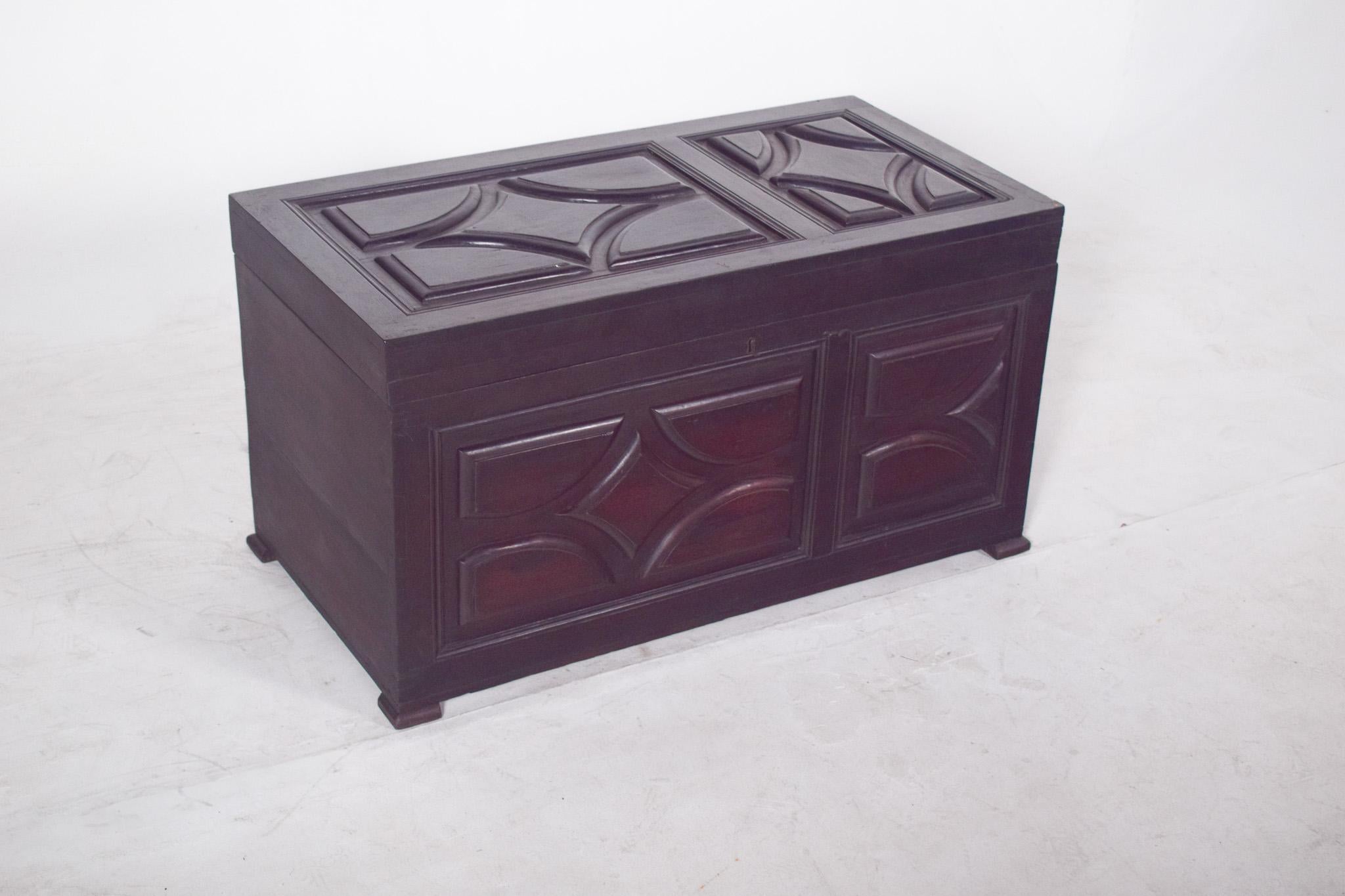 Cassapanca in legno imbottita. Con un coperchio a cerniera e una finitura scura, ha un design robusto e sobrio, che mette in risalto i pannelli pad sulla parte anteriore e superiore. I cuscini presentano dettagli geometrici in rilievo che aggiungono
