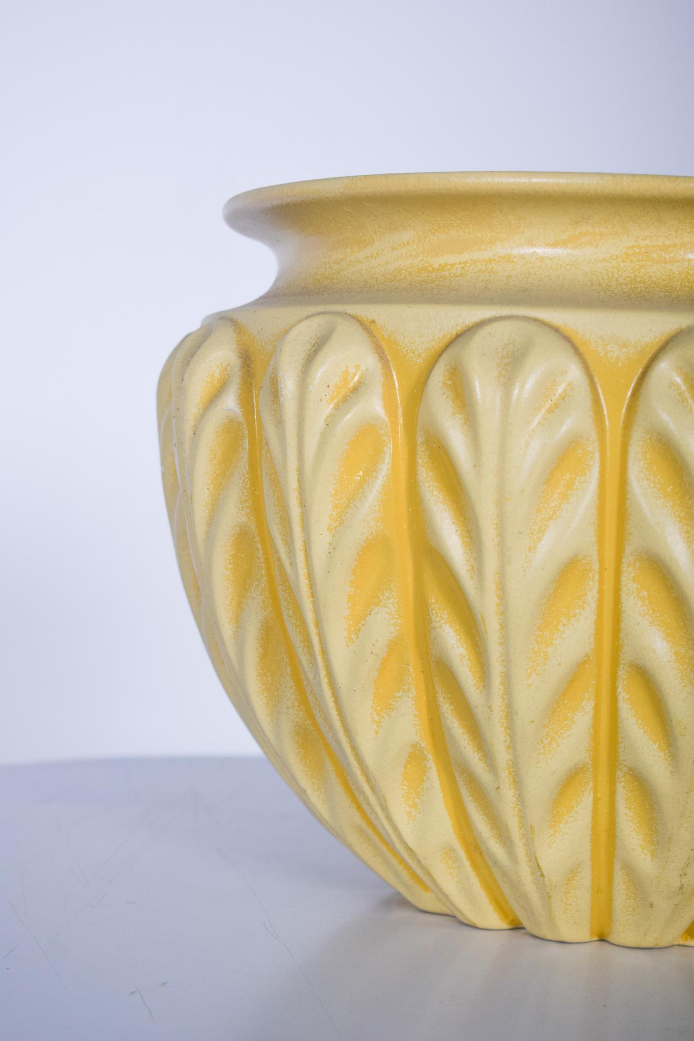 Cache pot en céramique jaune. La pièce présente un design arrondi et robuste, avec des détails en relief, qui sont des feuilles stylisées, réparties harmonieusement sur toute la surface. Cette texture élaborée crée un effet tridimensionnel, faisant