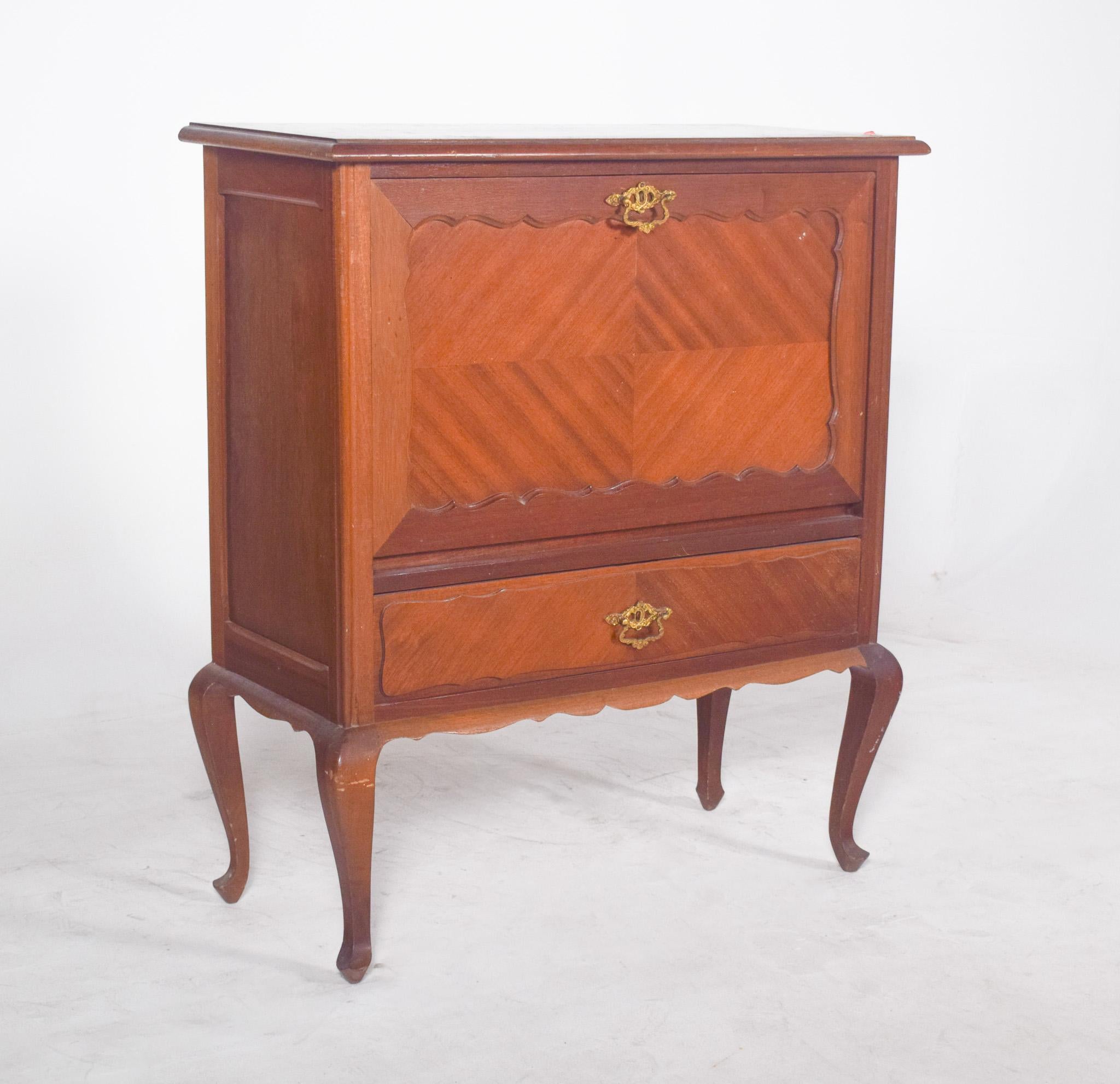 Mueble bar estilo Queen Anne. Marcada por la ligereza de sus formas y la elegancia de sus detalles, la pieza descansa sobre cuatro patas cabriolet, de líneas curvas que dan fluidez a la estructura y refuerzan su estética clásica.
La parte frontal