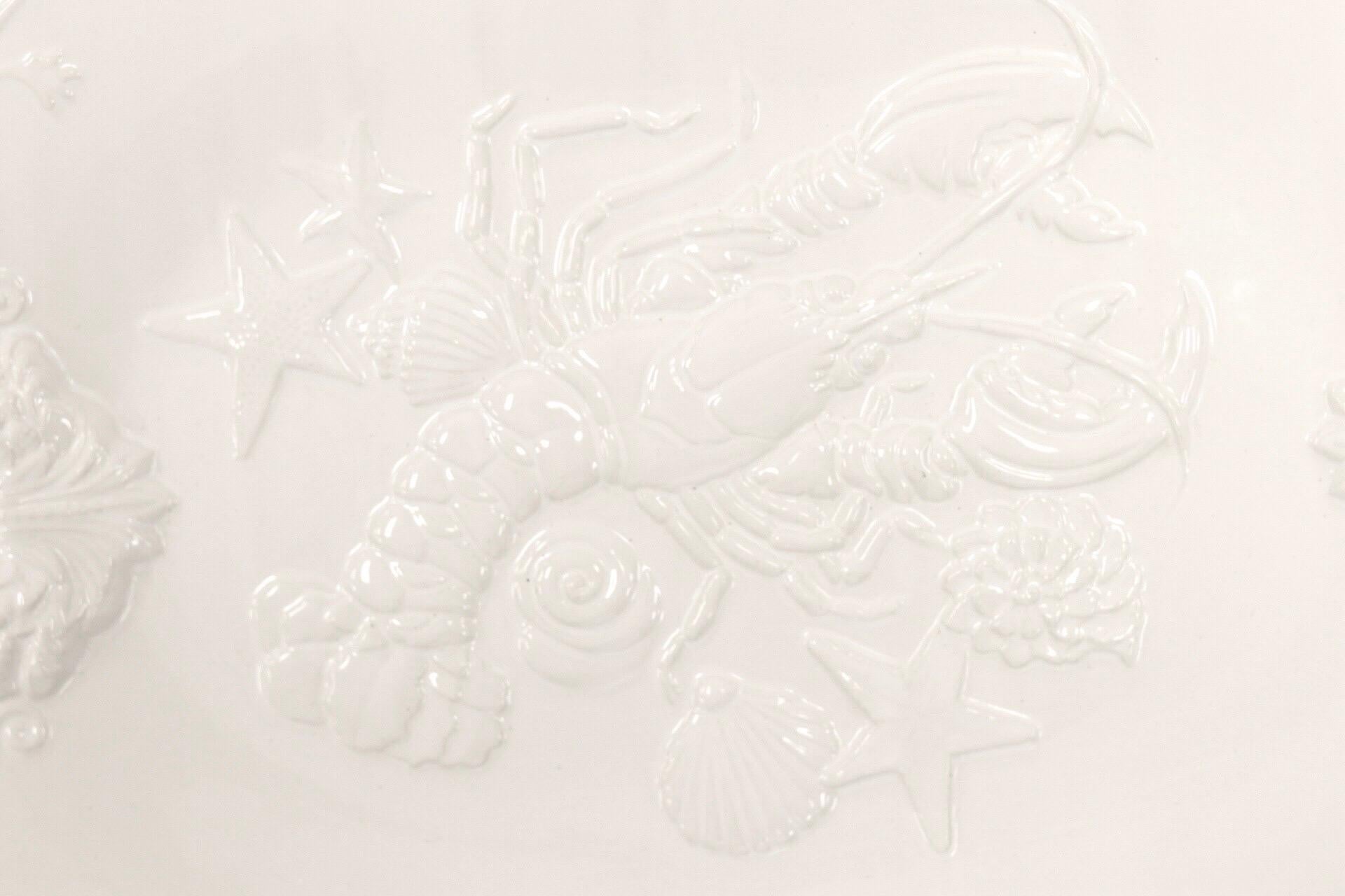 A white ceramic platter made by Sanor Ceramica of Portugal. Embossed with a coastal design, centered with a lobster amidst starfish and shells. Each end of the platter is embossed with a scallop shell, and the edge is decorated with trailing