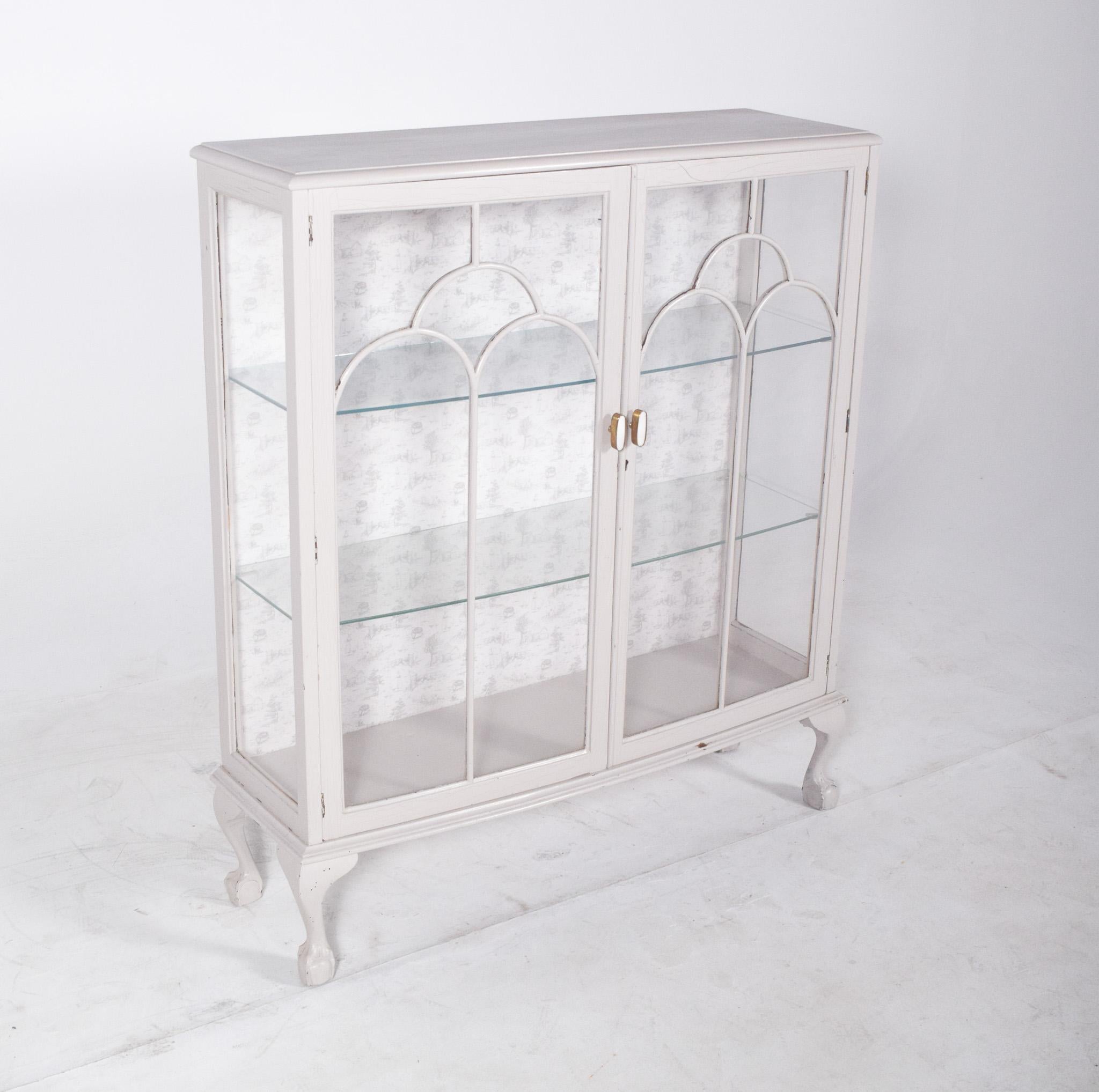 Vetrina dipinta di bianco. Dotata di porte e lati vetrati, presenta tre ripiani interni in vetro che garantiscono una buona visibilità e luce agli oggetti esposti. Le porte d'ingresso hanno maniglie dorate e cornici con archi decorativi, che